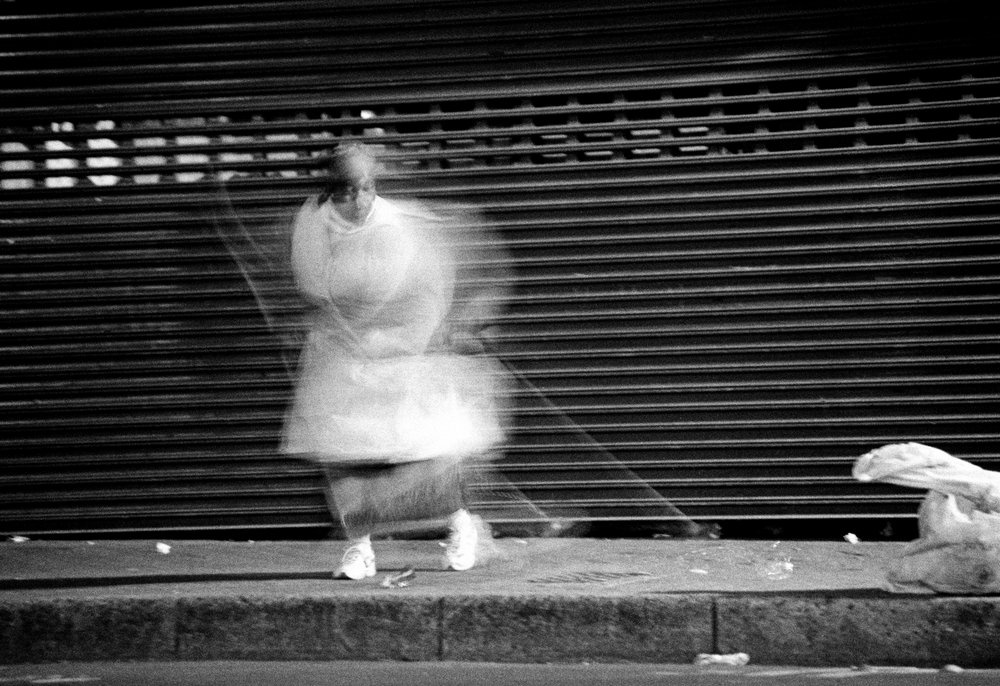 Sabelo Mlangeni's monochrome photographic print 'Invisible Woman II' shows a woman walking on a concrete sidewalk.
