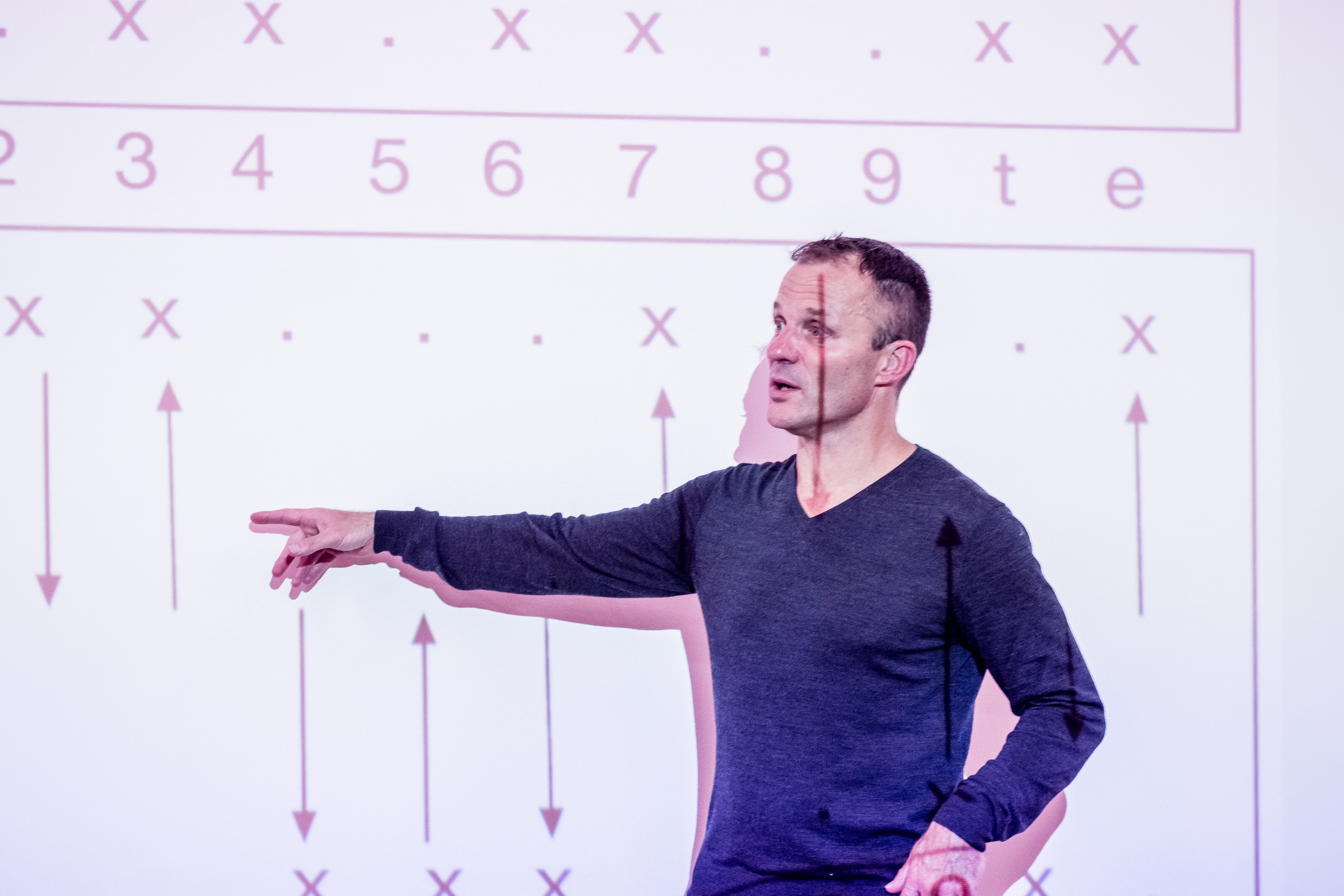 Event photograph from the ‘Algorithmic Temporalities of the Neocolony: the Case of African Mathematics’ event on A4’s top floor. Professor Martin Scherzinger stands in front of a white wall, pointing at an area of the diagram projected onto the wall behind him.
