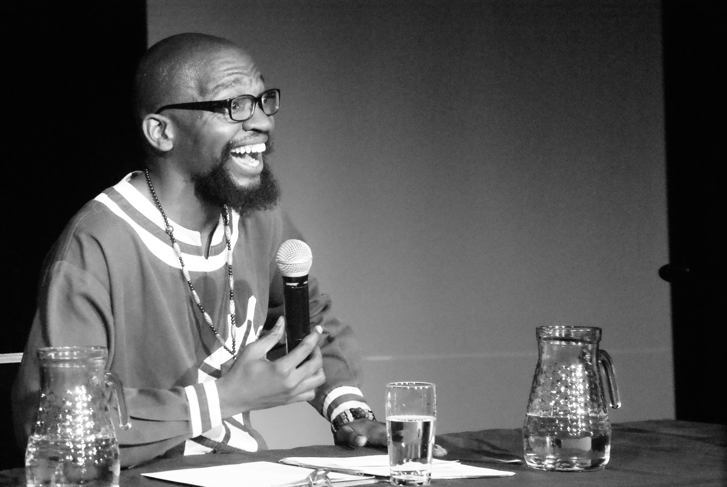 Monochrome event photograph from Jefferson Bobs Tshabalala’s residency on A4’s top floor that shows the artist seated at a table with a microphone.
