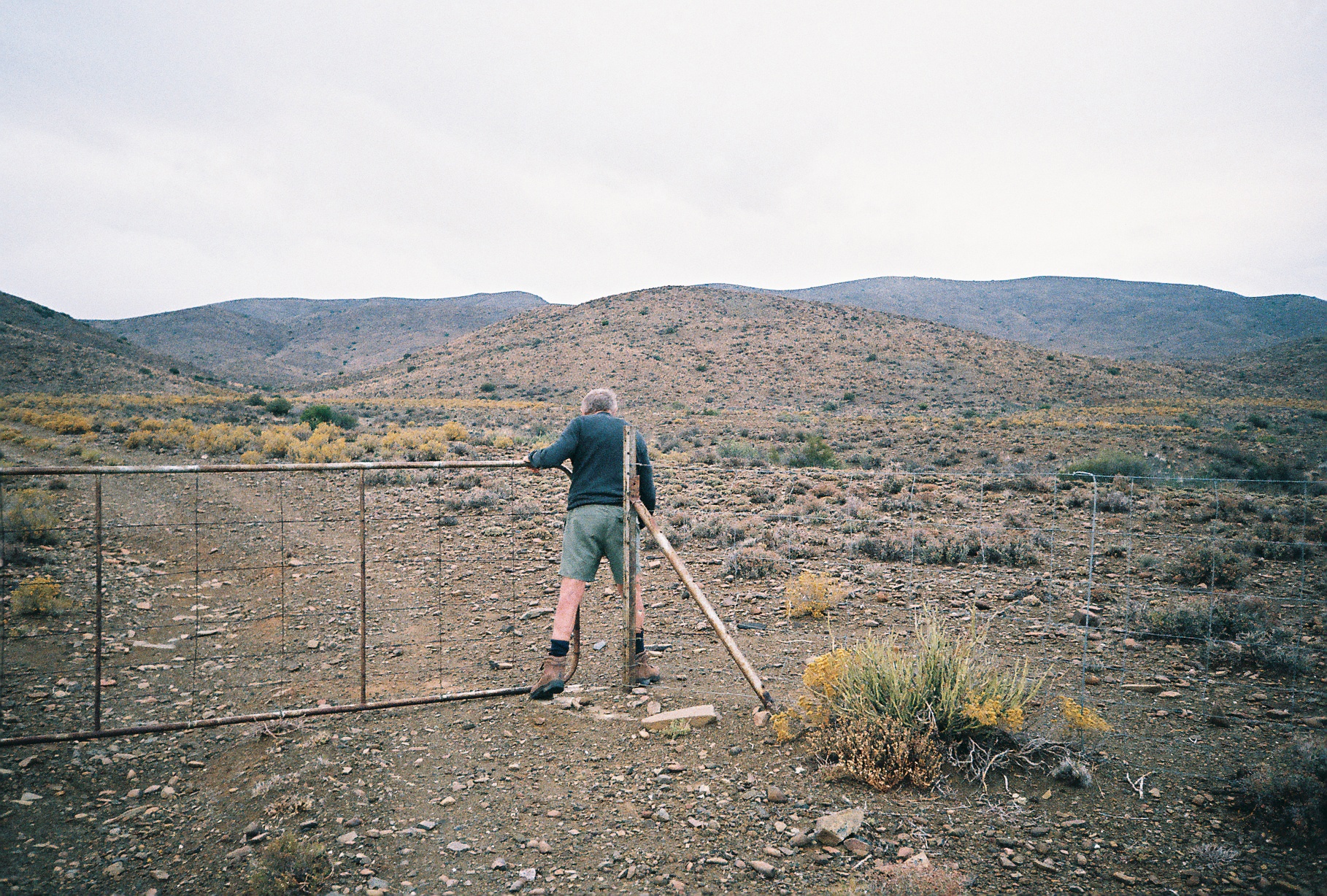 Still frame from the Goldblatt documentary directed by Daniel Zimbler shows David Goldblatt walking through a gate in a wire fence.
