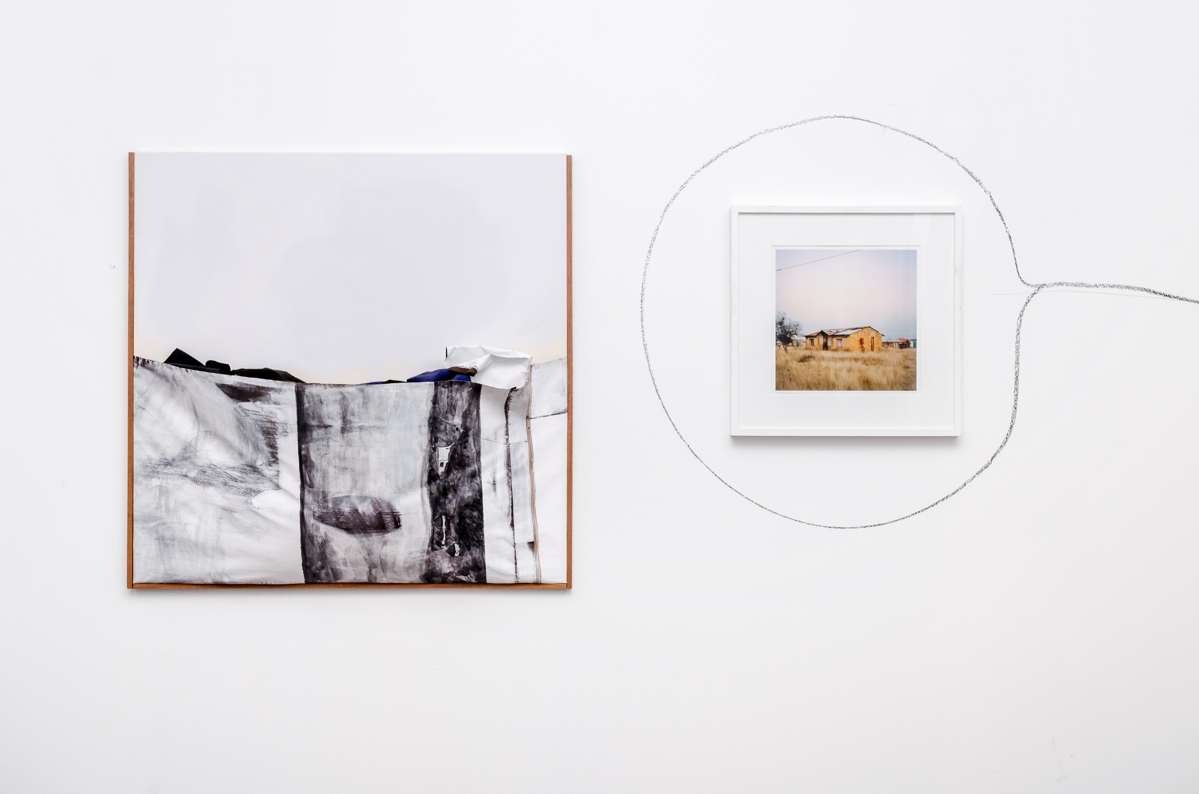 Installation photograph from the ‘Between Two or More Worlds’ exhibition at the 2016/2017 A4 Office. On the left, Gerda Scheepers’ acrylic painting ‘Pondoland pocket’ is mounted on the white wall. On the right, Thabiso Sekgala’s photograph ‘Semotlhase, former Bophuthatswana’ is mounted on the white wall and encircled with charcoal.
