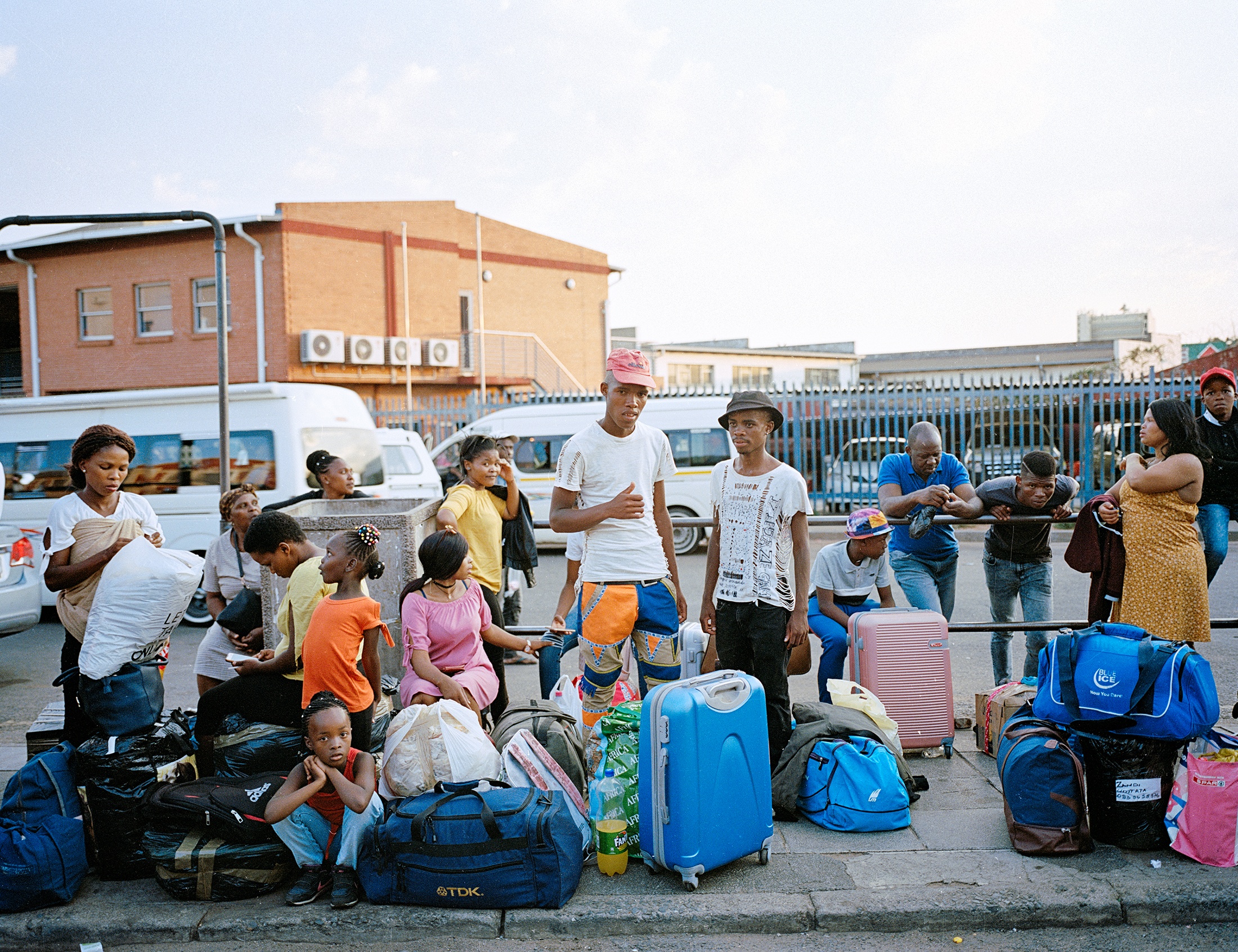 Lindokuhle Sobekwa's photograph 'Germiston bus stop' depicts a group of people with luggage on a roadside pavement.
