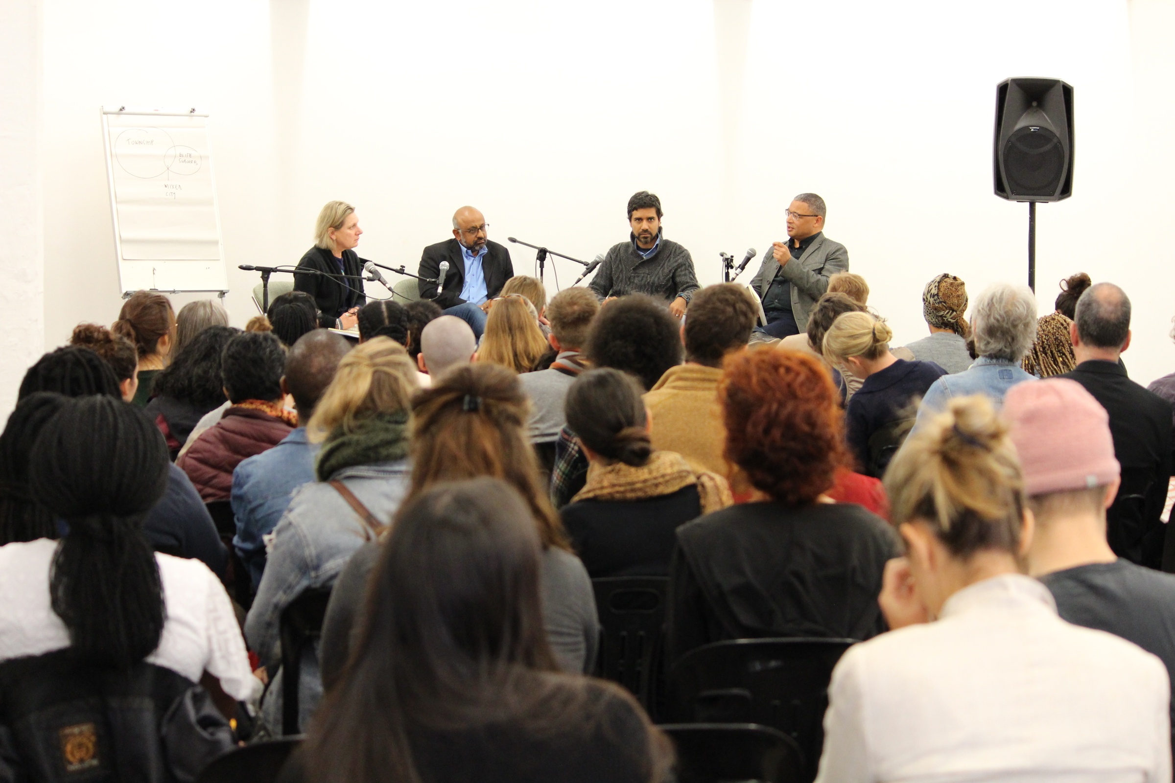 Event photograph from the 2017 rendition of the Open Book literary festival on A4’s ground floor. At the front, attendees are seated. At the back, Pippa Green, Premesh Lalu, Adi Kumar and Edgar Pieterse are seated with standing microphones.
