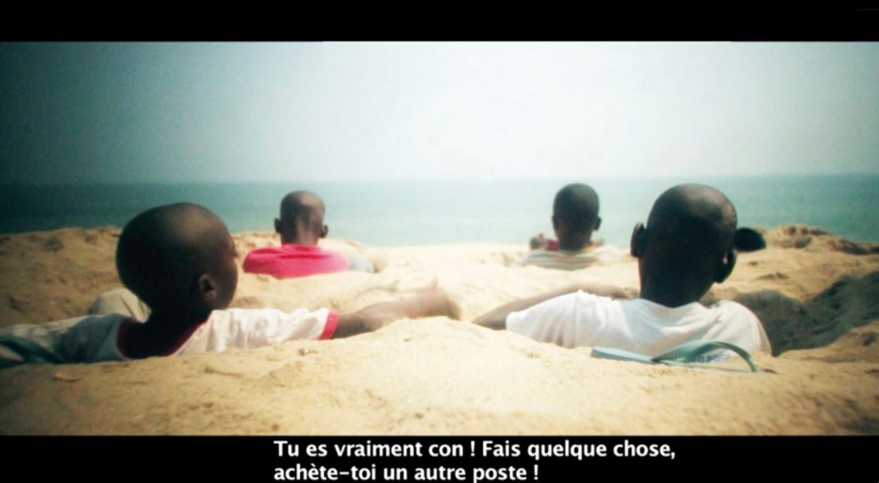 A still frame from Binelde Hycran’s video work ‘Cambeck’ that shows four children sitting in holes dug in beach sand to resemble a vehicle with the subtitles reading “Tu es vraiment con ! Fais quelque chose, achète–toi un autre poste !”
