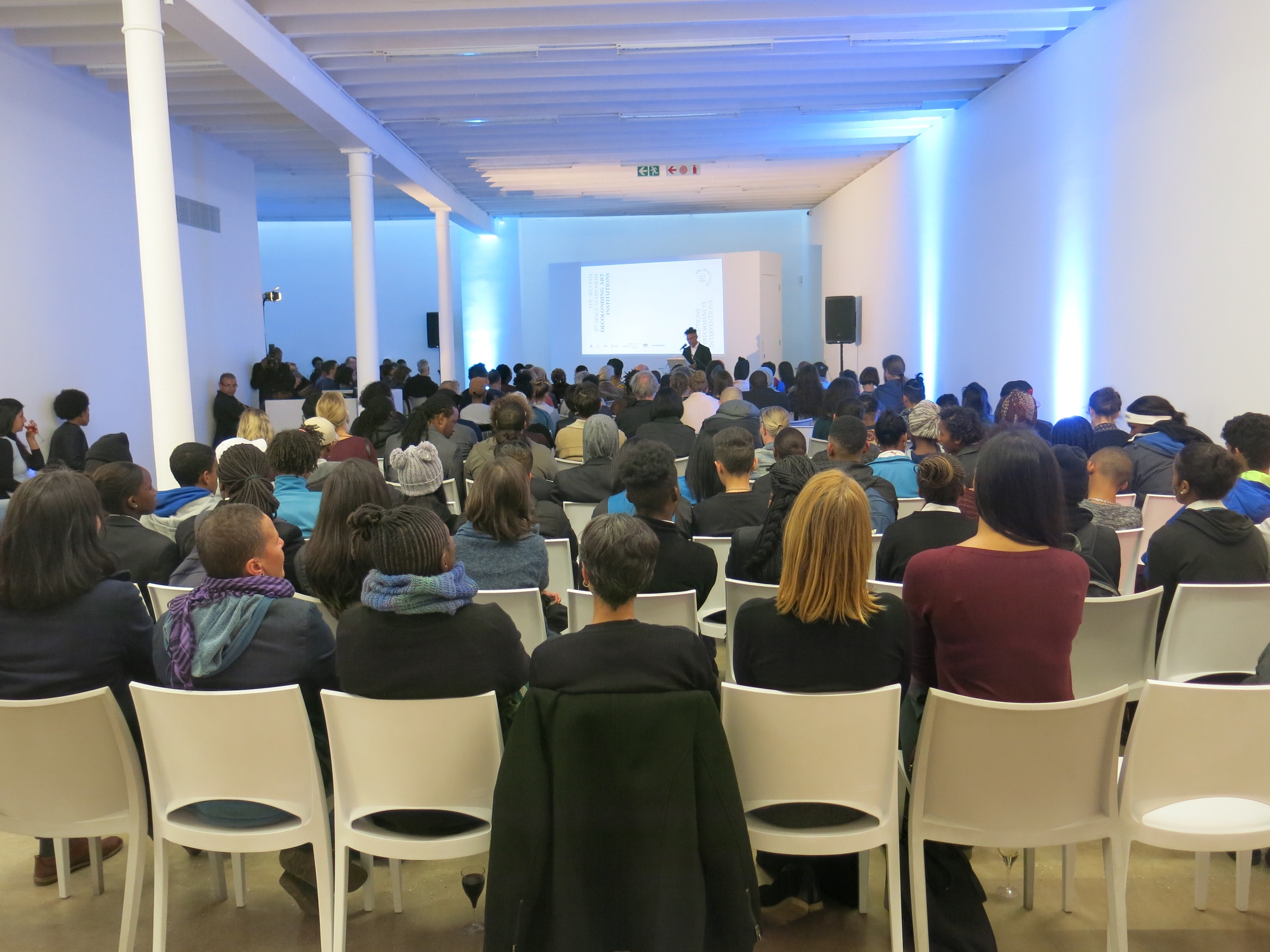 Event photograph from the ‘3rd Space Symposium’ in A4’s gallery. At the front, seated attendees fill most of the gallery space. At the back, a speaker stands before a wall projection.
