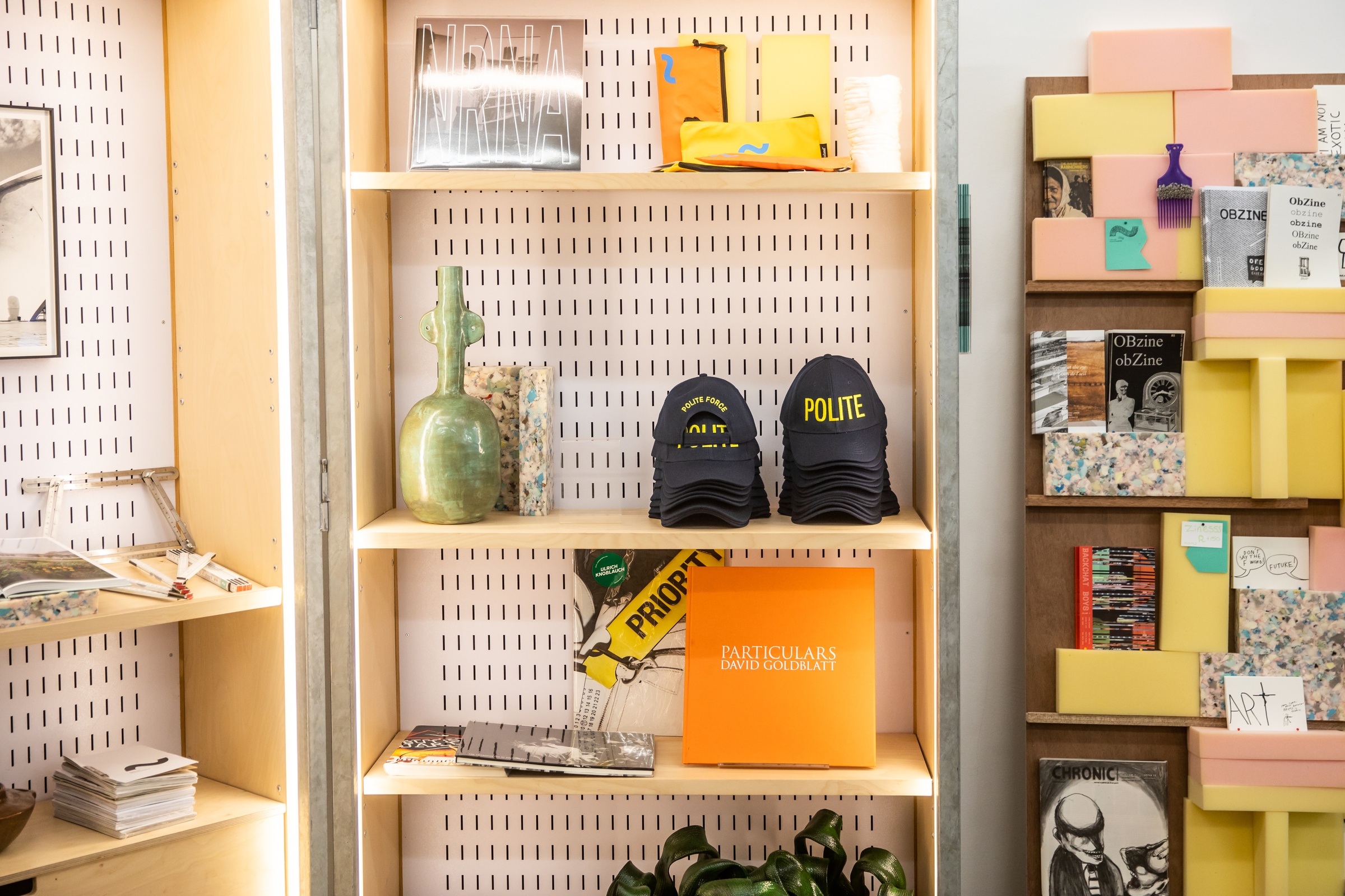 Event photograph from the ‘Hello proto~’ launch of A4’s Proto~ museum shop. In the middle, a cabinet is lined with various artworks and ephemera. On the top shelf, Never Read the News Alone’s vinyl EP ‘Kalahari Elegy.’ To the middle, Christian Nerf’s ‘Polite Force’ caps. To the bottom, David Goldblatt’s photo-book ‘Particulars.’
