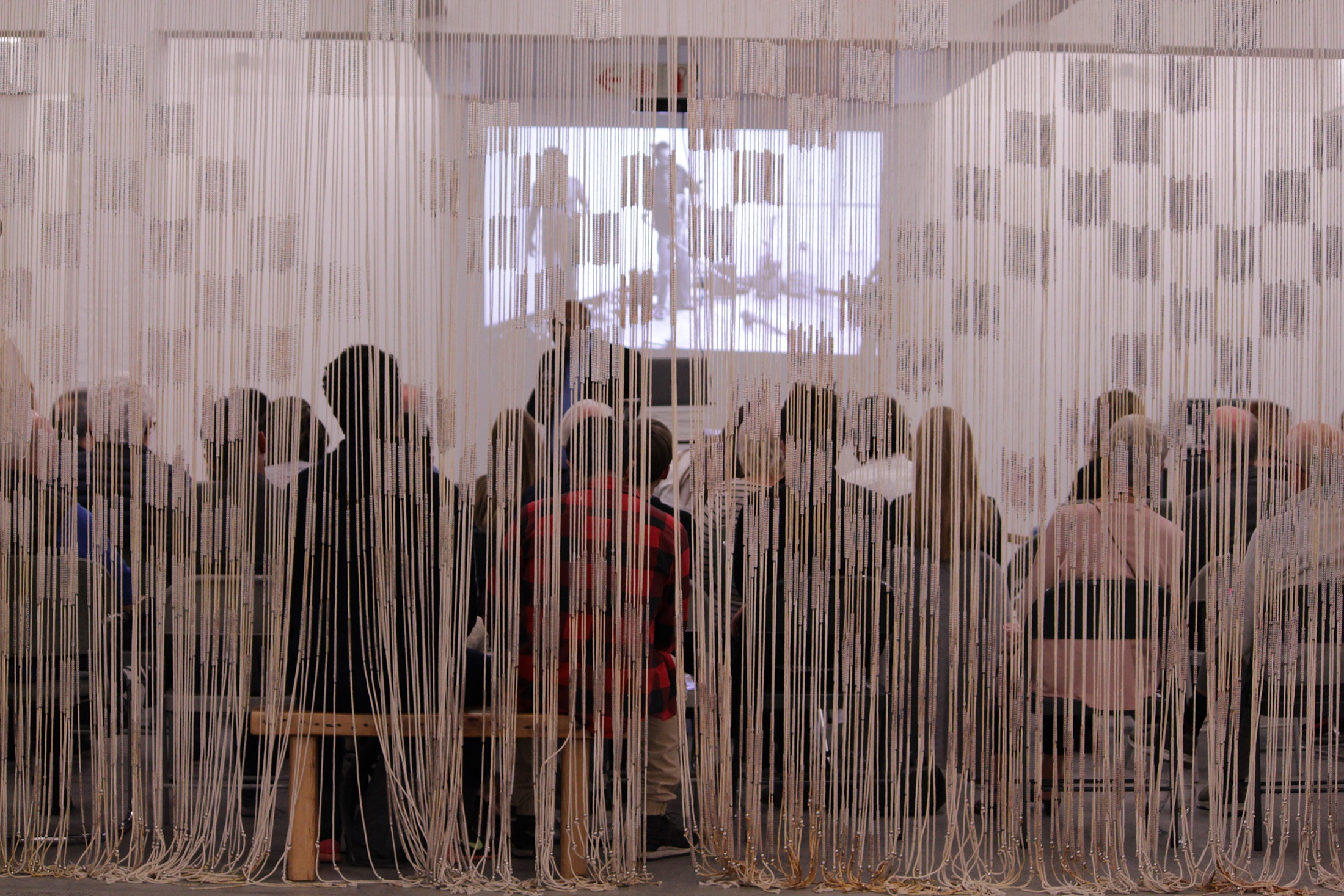 Event photograph from the book launch of Kim Gurney’s ‘August House is Dead, Long Live August House!’ on A4’s ground floor. At the front, Igshaan Adams’ mixed media tapestry ‘Al asma ul Husna’ is suspended from the ceiling. At the back, attendees sit in rows in front of the speakers and a projection on the far wall.
