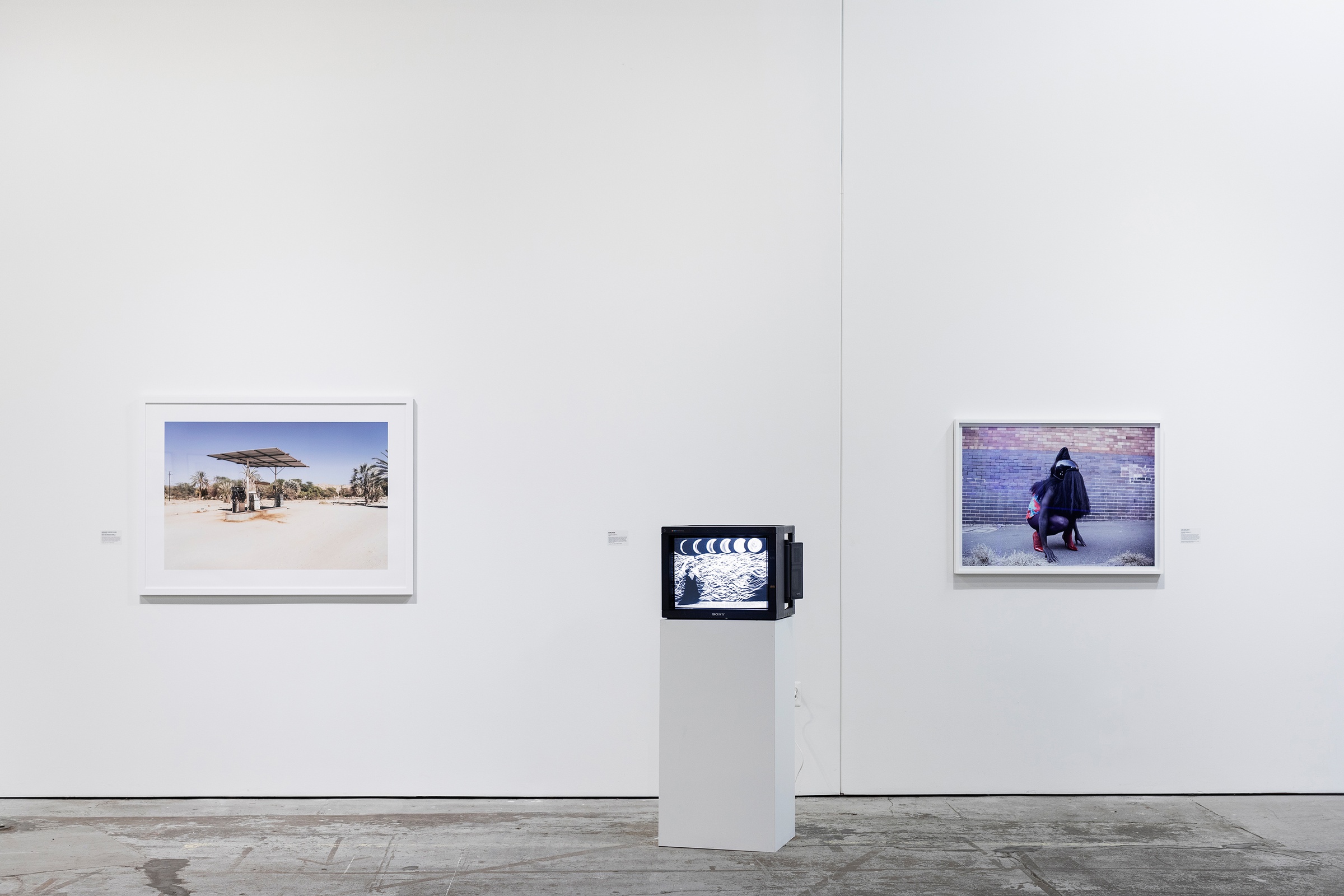 Installation photograph from ‘Crossing Night: Regional Identities x Global Context’ exhibition at the Museum of Contemporary Art Detroit. On the left, Margaret Courtney Clarke’s photograph ‘Petrol Pump, Sesfontein, Namibia’ is mounted on the wall. In the middle, Robin Rhode’s video ‘The Moon is Asleep’ is displayed on a screen sitting on a plinth. On the right, Athi-Patra Riga’s photograph ‘The Naivety of Beiruth’ is mounted on the gallery wall.
