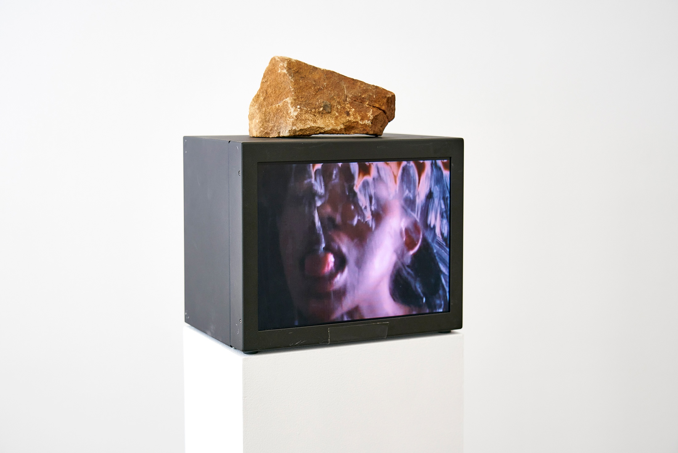 Installation photograph that shows Dineo Seshee Bopape’s video work ‘a love supreme’ playing on a video box that rests on a white plinth with a rock sitting on top of it.
