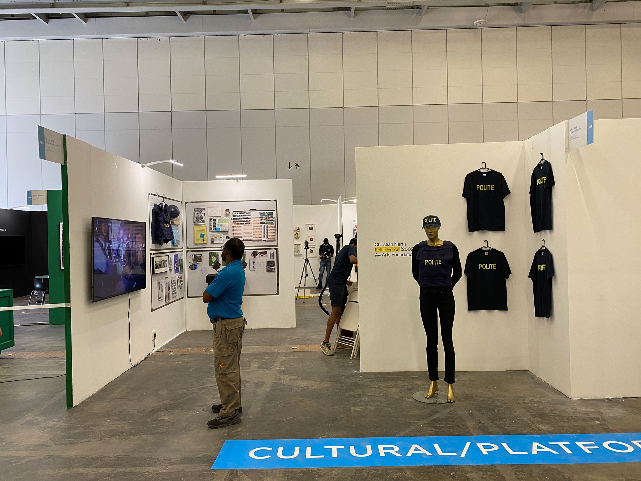 Event photograph from A4’s booth at the 2022 Investec Cape Town Art Fair. On the right, a mannikin dressed in Christian Nerf’s ‘Polite Force’ riot gear stands before a display of ‘Polite Force’ t-shirts. On the left, a video screen, photographic prints and objects line the booth wall, with an attendee looking at them.
