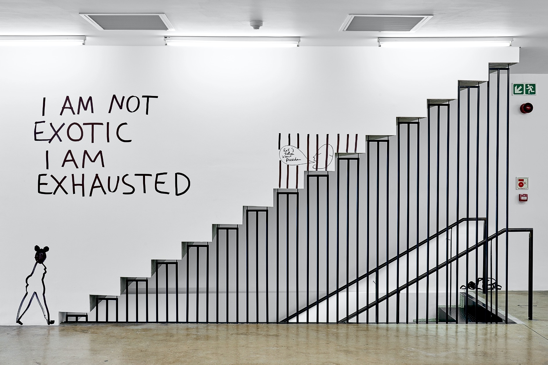 Installation photograph from Dan Perjovschi’s ‘The Black and White Cape Town Report’ exhibition in A4’s Gallery that depicts a sidelong view of a staircase. On the right, the descending stairway is visible through black guardrails. On the left, the white wall features black pen marker text that reads ‘I AM NOT EXOTIC, I AM EXHAUSTED’, with a humanoid figure below it.
