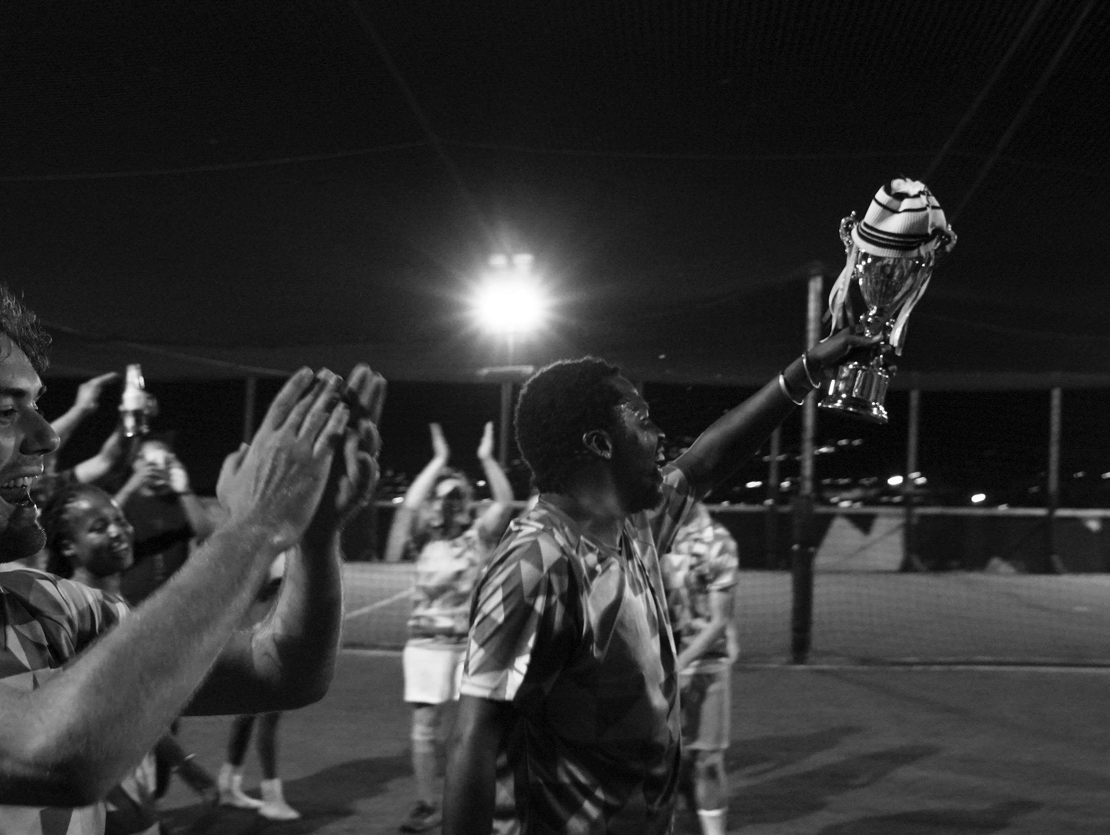 Monochrome event photograph from the 2023 edition of Exhibition Match depicts the winners of a soccer match holding a trophy on a lit rooftop field at night.
