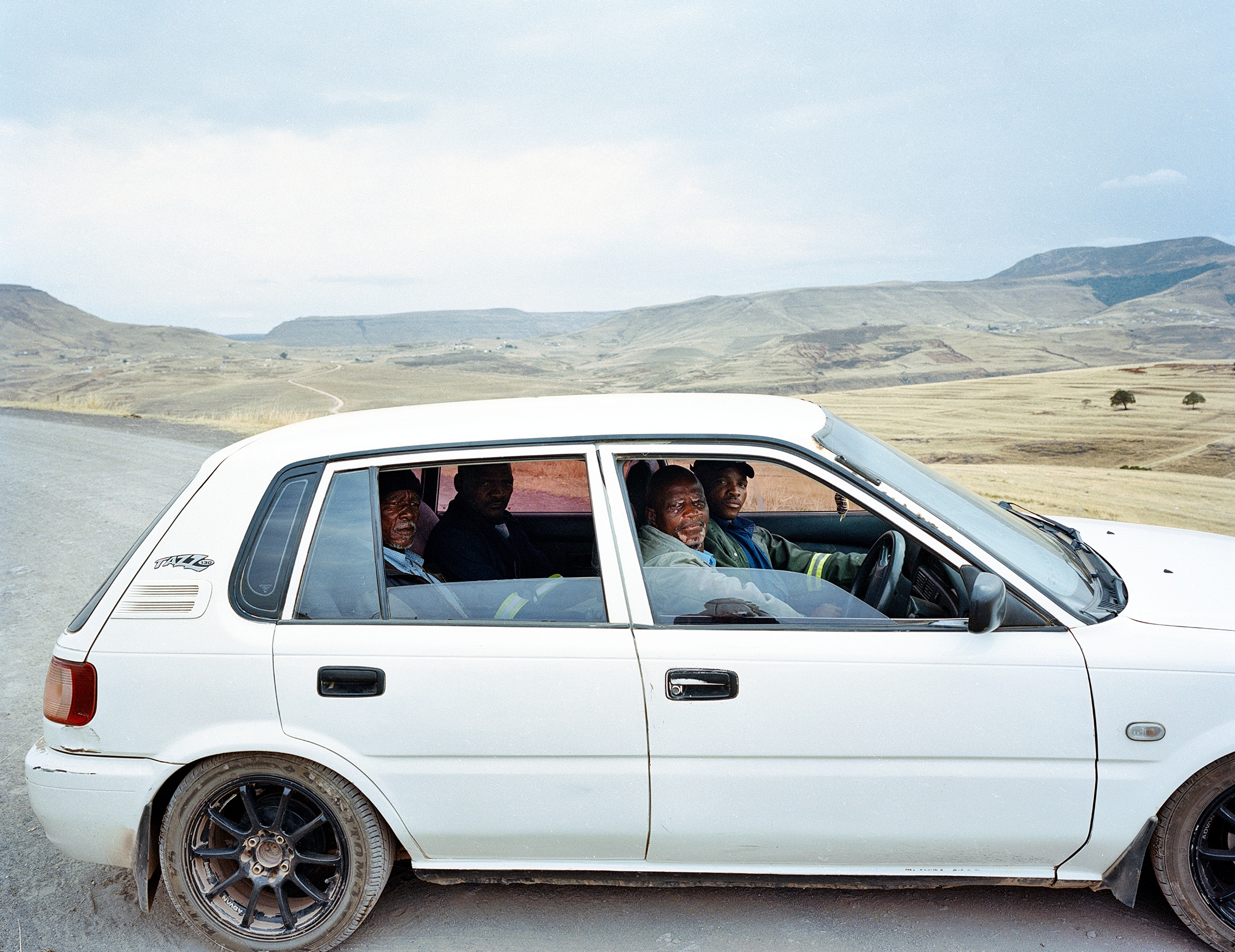 Lindokuhle Sobekwa's photograph 'Ntasbasgogo Qumbu' depicts four individuals sitting in a white car on a tarred road at the front, with grassy plateaus at the back.  
