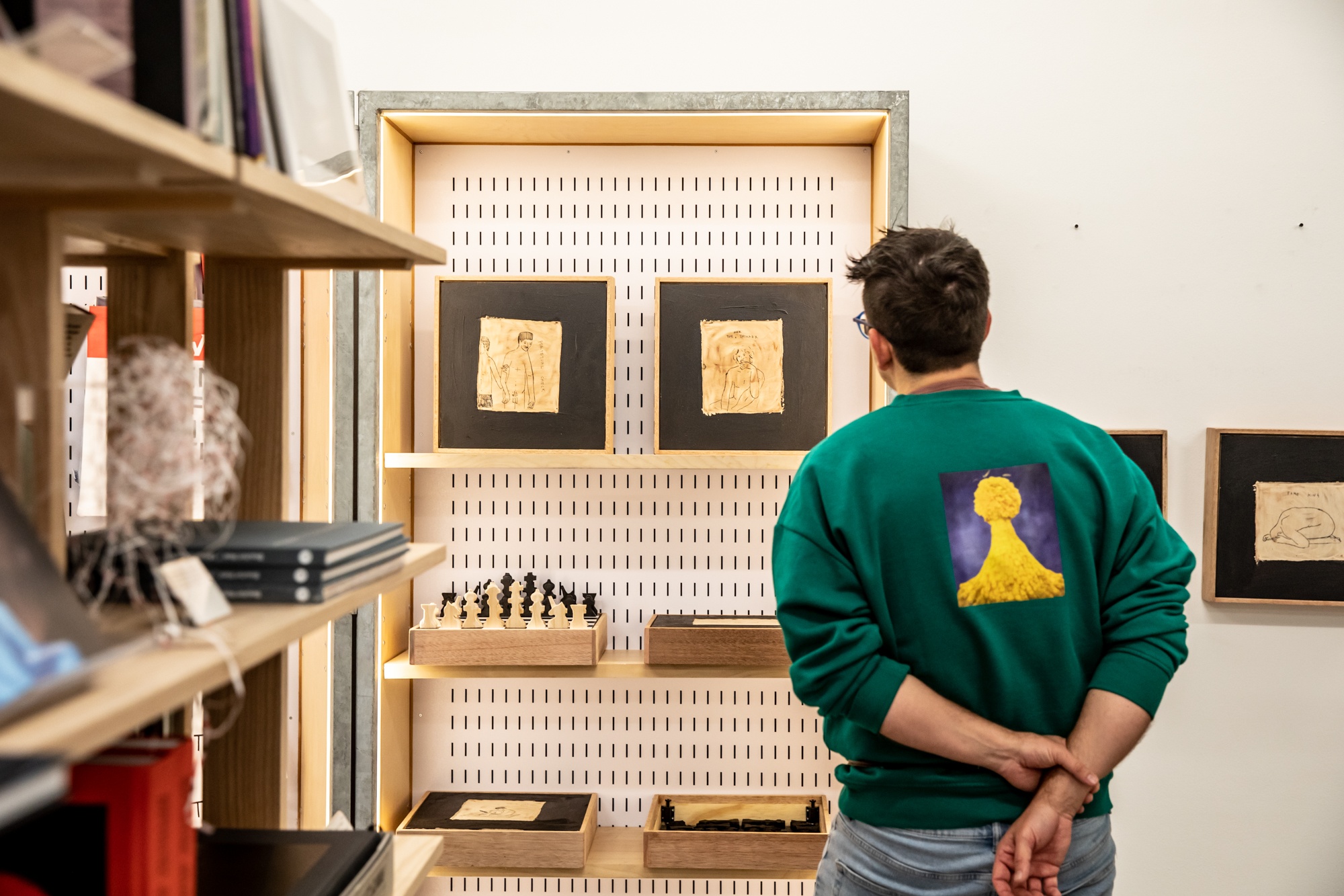 Event photograph from the launch of Brett Seiler’s chess set in A4’s Proto museum shop. A display case against the shop wall holds multiple wooden chess sets with paintings on the bottom of each set. An attendee faces the display with their back to the photographer.
