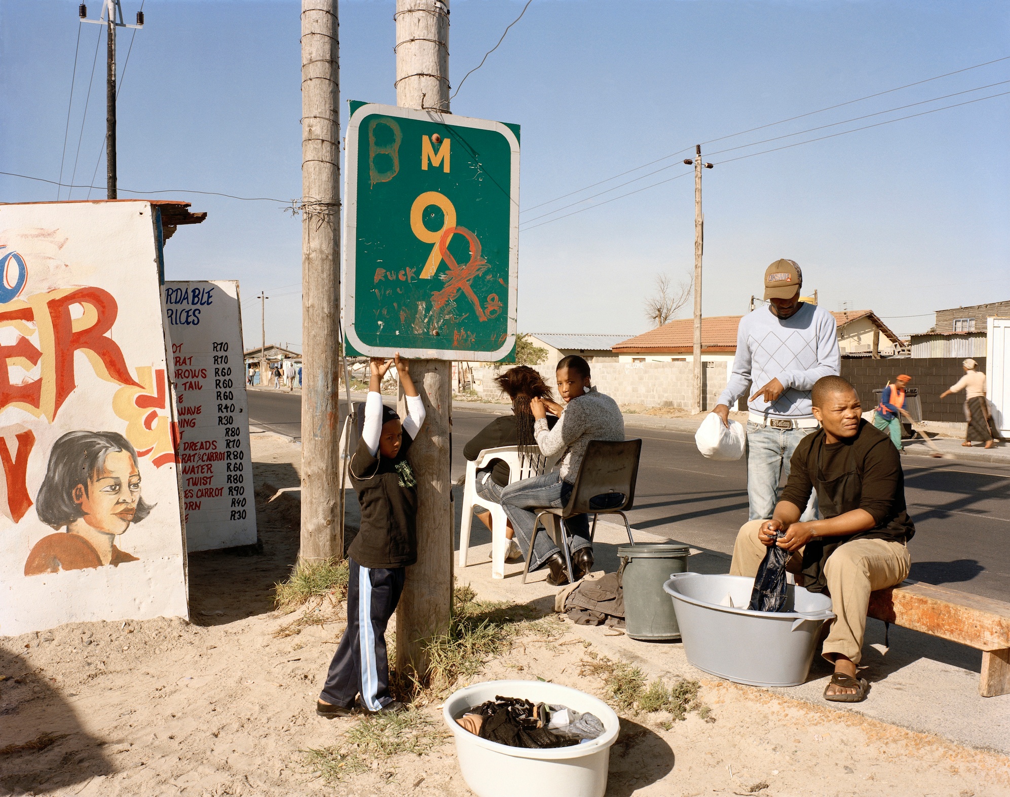 David Goldblatt's photograph 'At Kevin Kwanele’s Takwaito Barber. Lansdowne Road, Khayelitsha, Cape Town in the time of AIDS' shows individuals seated on a sidewalk. At the back, an individual is braiding another's hair. At the front, an individual is washing clothes in a plastic bucket.
