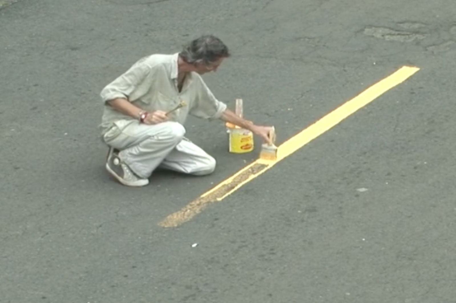 A still frame from Francis Alÿs’ video work ‘Painting/Retoque’ shows a person crouched on a tarred road repainting a faded road marking with yellow paint.

