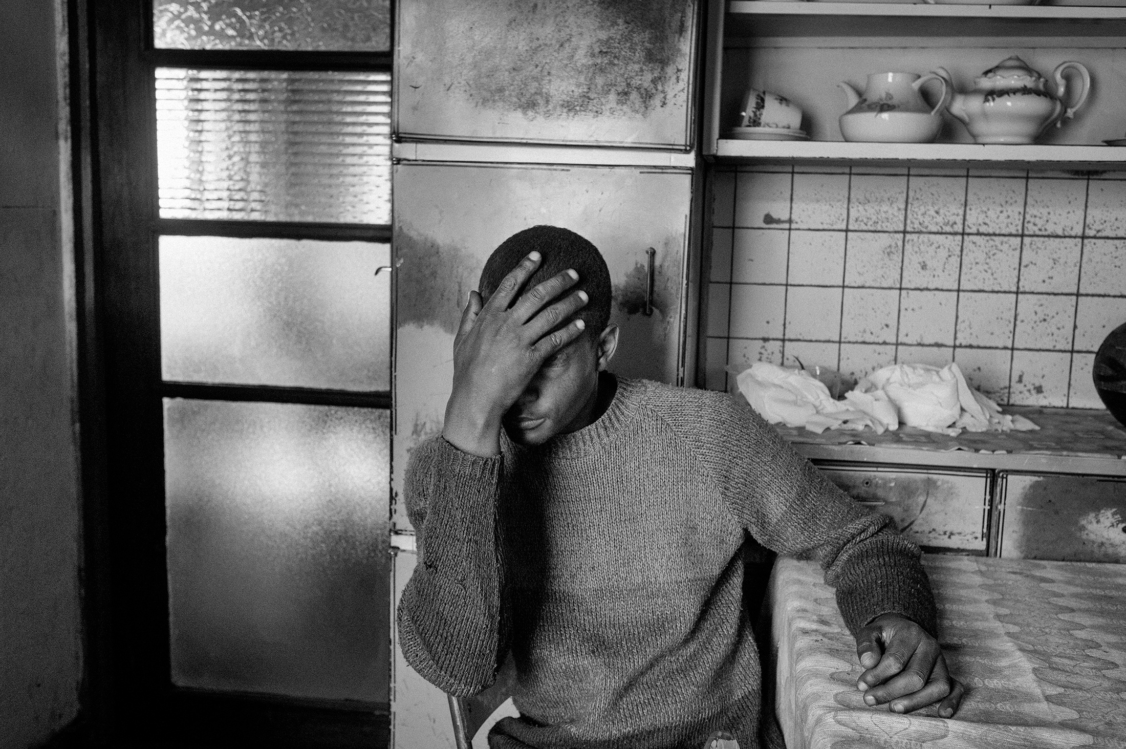 Lindokuhle Sobekwa's monochrome photograph 'Mzwandile at home after coming from the rehab centre' shows an individual sitting at a kitchen table with their their hand on their head.
