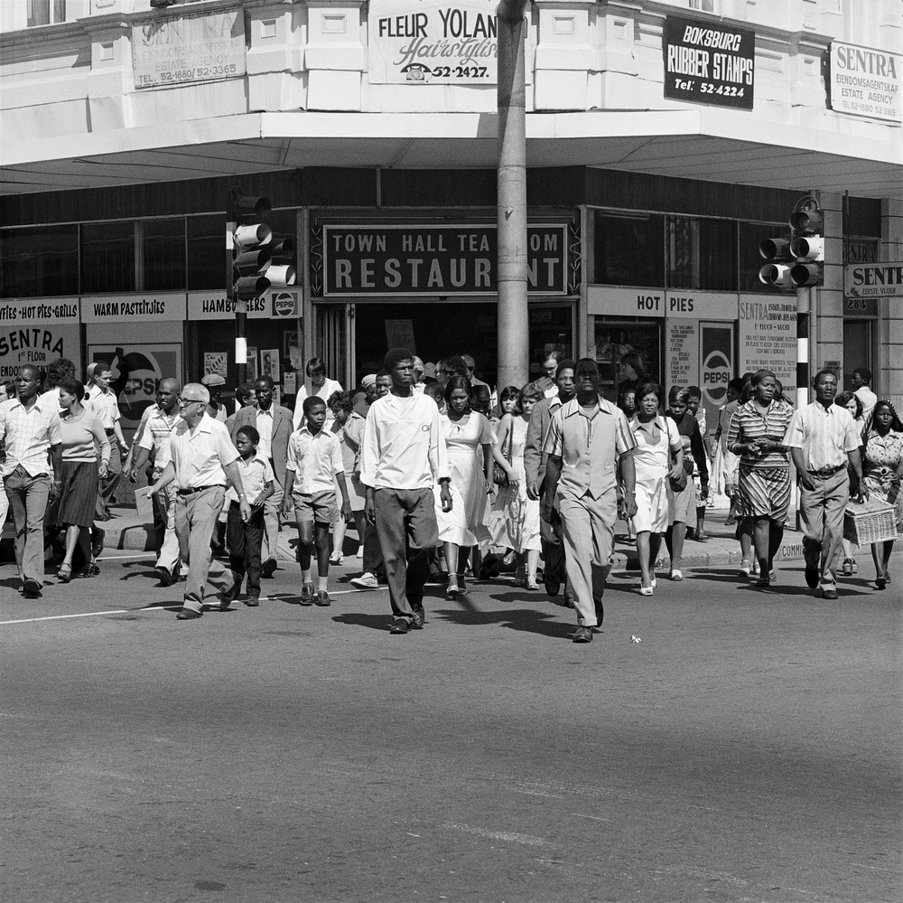 David Goldblatt's black-and-white photograph 'Saturday morning at the corner of Commissioner and Trichardt Streets, Boksburg' shows a crowd walking across a road in front of storefronts. 
