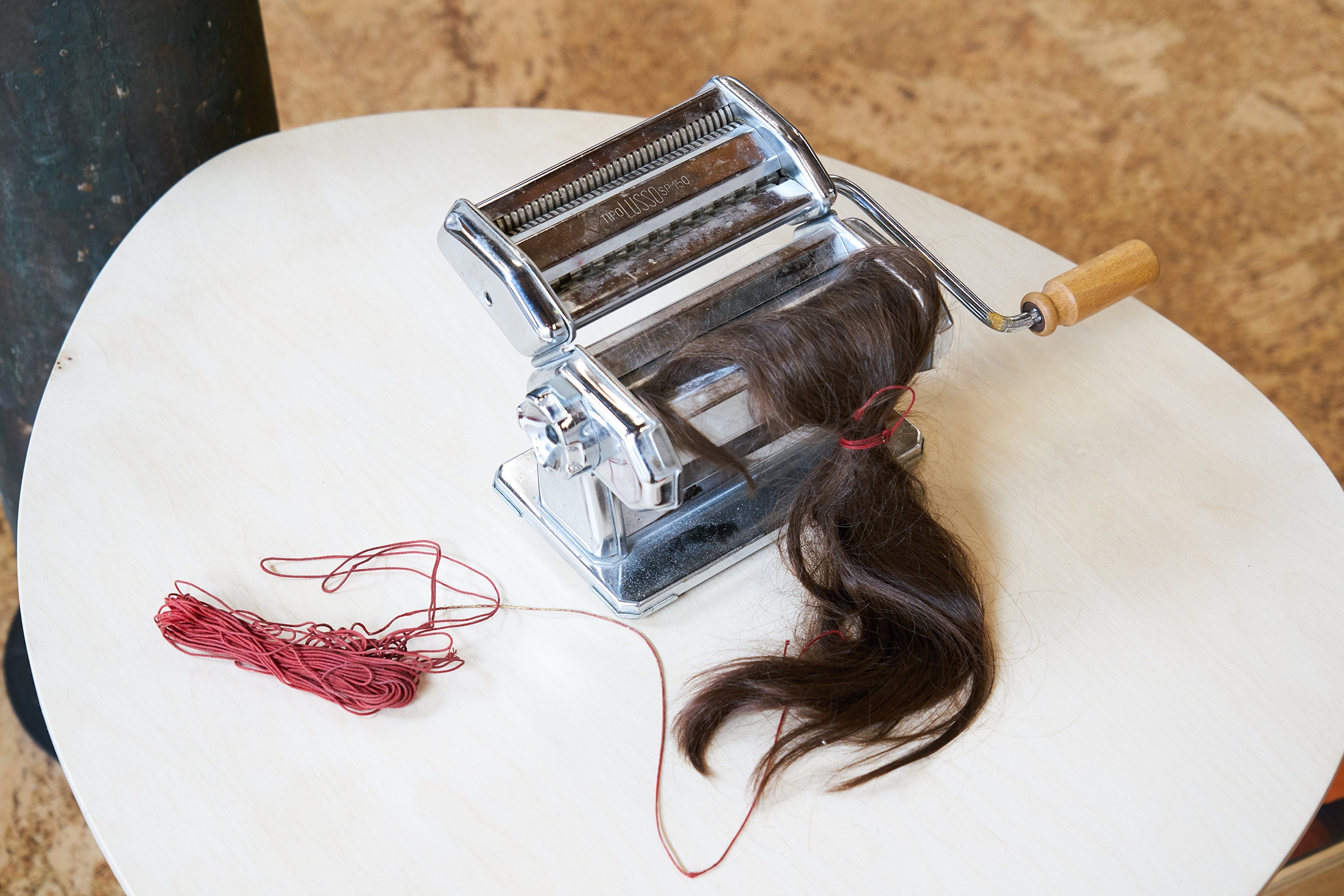 Process photograph from Christian Nerf’s residency on A4’s top floor. A pasta machine sits on a white table, with brown hair tied with red string rolled into the machine.
