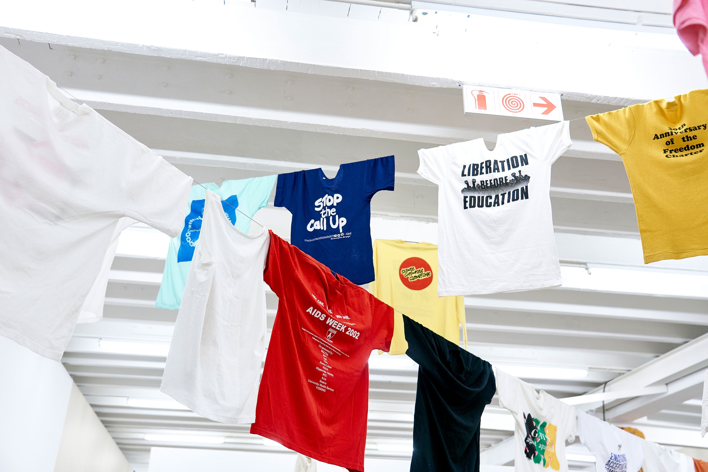 Installation photograph of the Common exhibition. Above, T-shirts with slogans from the GALA Queer Archive and SAHA are suspended on crossing lines. One white T-shirt features images of raised fists and reads “Liberation before Education.”
