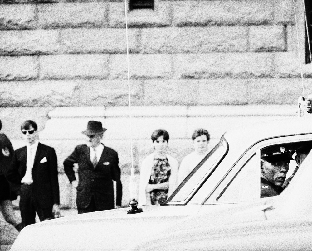 David Goldblatt's black and white photograph 'Policeman in a squad car on Church Square, Pretoria (Tshwane), Transvaal (Gauteng)' shows a man in a car with a row of people standing behind.
