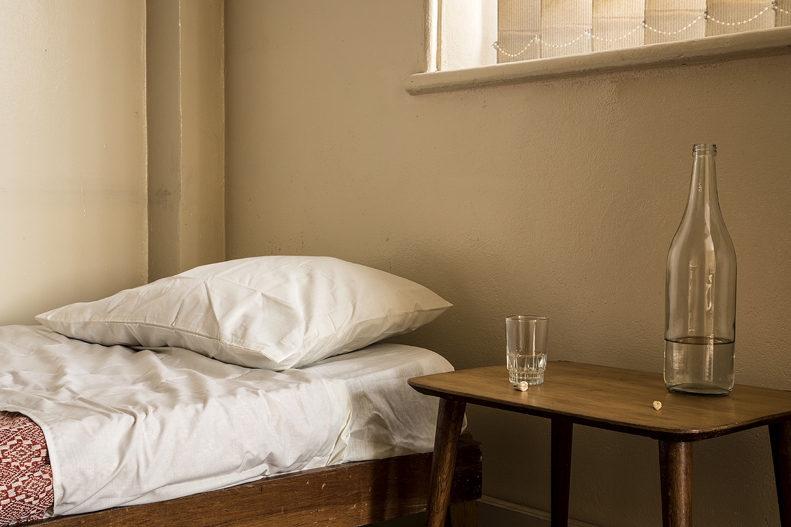 Installation photograph from Gain Maria Tosatti’s offsite exhibition ‘My Hart is so Leeg soon ’n Spieël (My Heart is a Void, the Void is a Mirror)’. On the left, a single bed with a pillow. On the right, a wooden side table with glass bottle, a drinking glass and two teeth.
