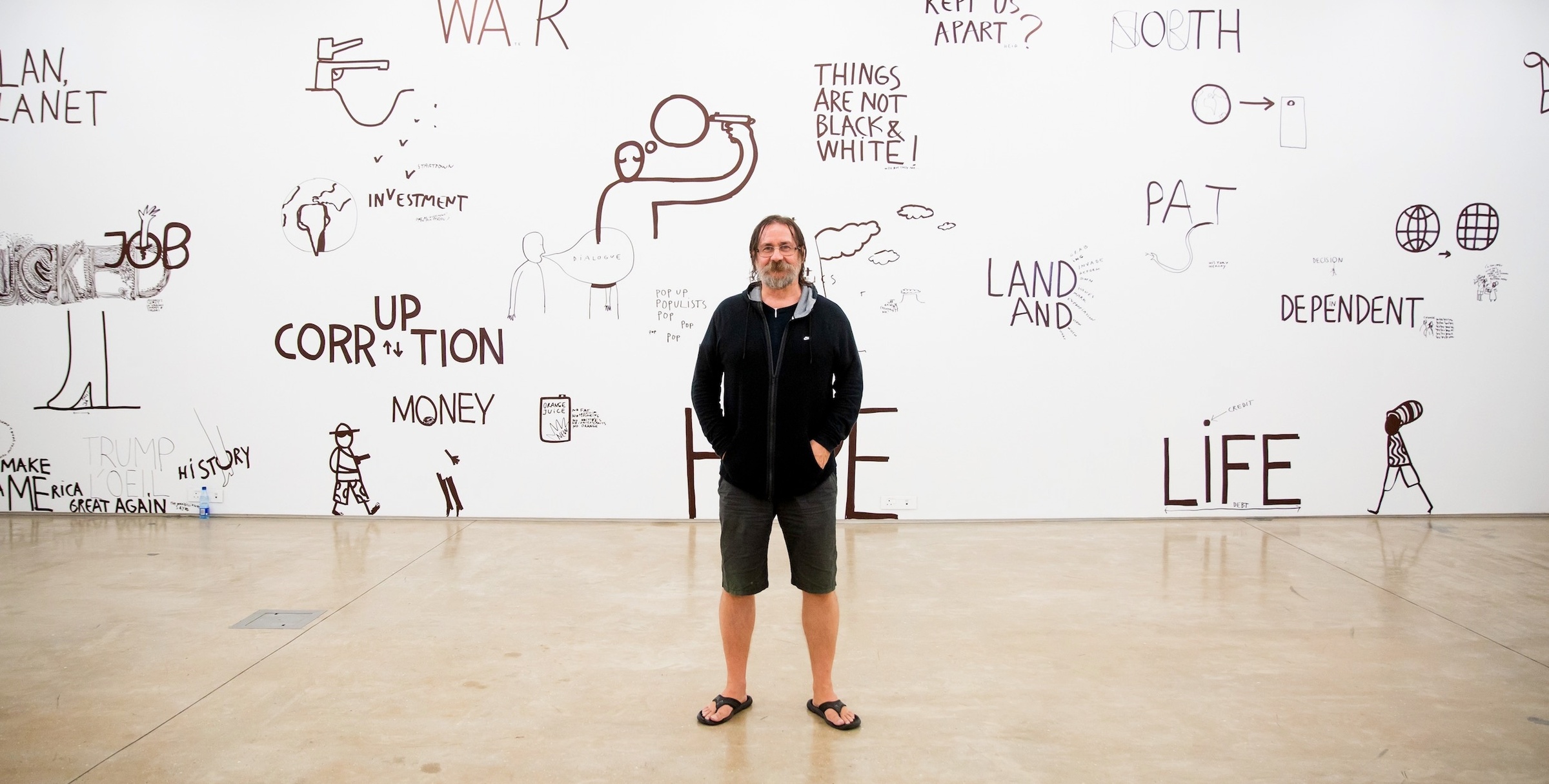 Event photograph from Dan Perjovschi’s presentation in A4’s Gallery. At the front, Perjovschi stands in the middle of the frame. At the back, a white wall covered in black felt pen marker drawings from his ‘Black & White Cape Town Report’ exhibition.
