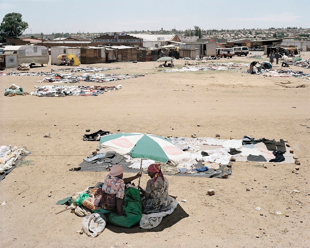 David Goldblatt's photograph 'Freedom Square: here, in the time of apartheid, on 26 June 1955, under harassment by the police, some 3000 people of all races, from all over South Africa, gathered in a Congress of the People and adopted the Freedom Charter, a template for the governance of a non-racial, democratic South Africa. The Charter became the basis of South Africa’s democratic constitution. Kliptown, Soweto, Johannesburg'. At the front, two individuals seated under an umbrella on sandy plain. At the back, various buildings make up the border of a settlement.
