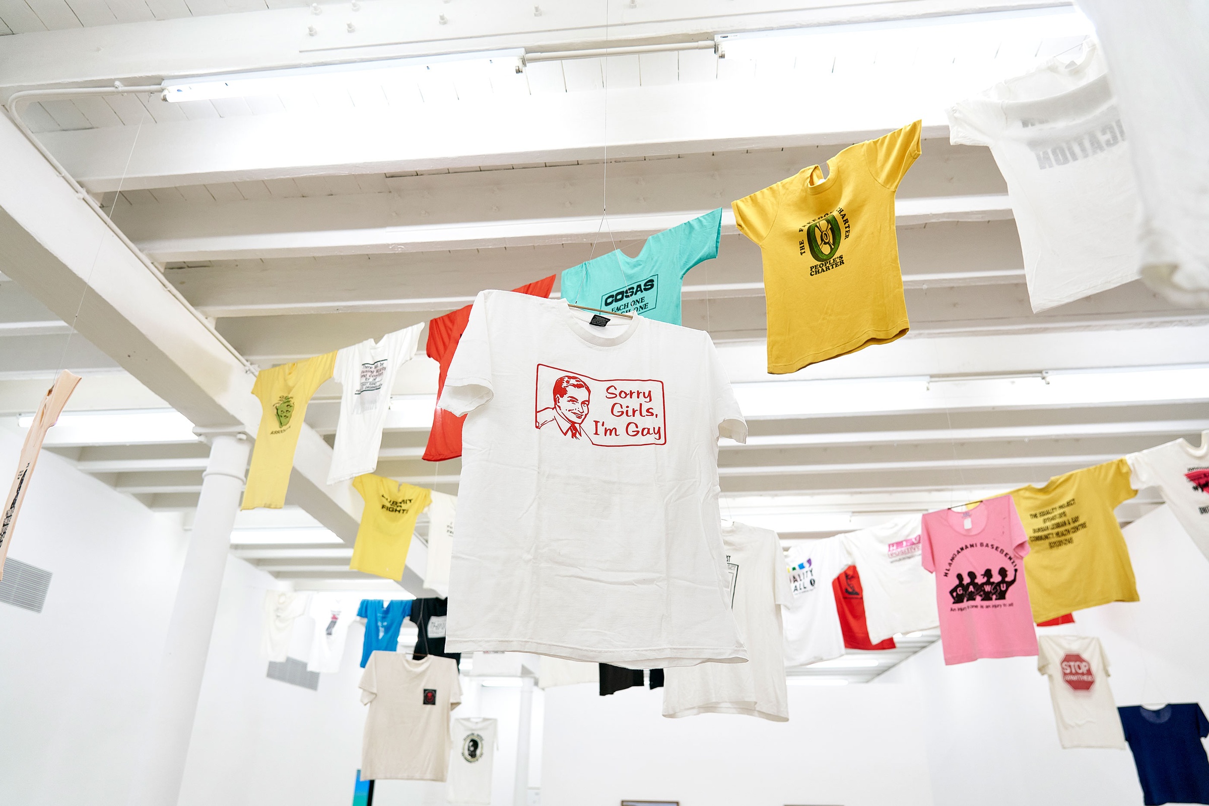 Installation photograph of the Common exhibition. Above, T-shirts with slogans from the GALA Queer Archive and SAHA are suspended on crossing lines. One white T-shirt features an image of a man’s smiling face and reads "Sorry Girls, I’m Gay.”
