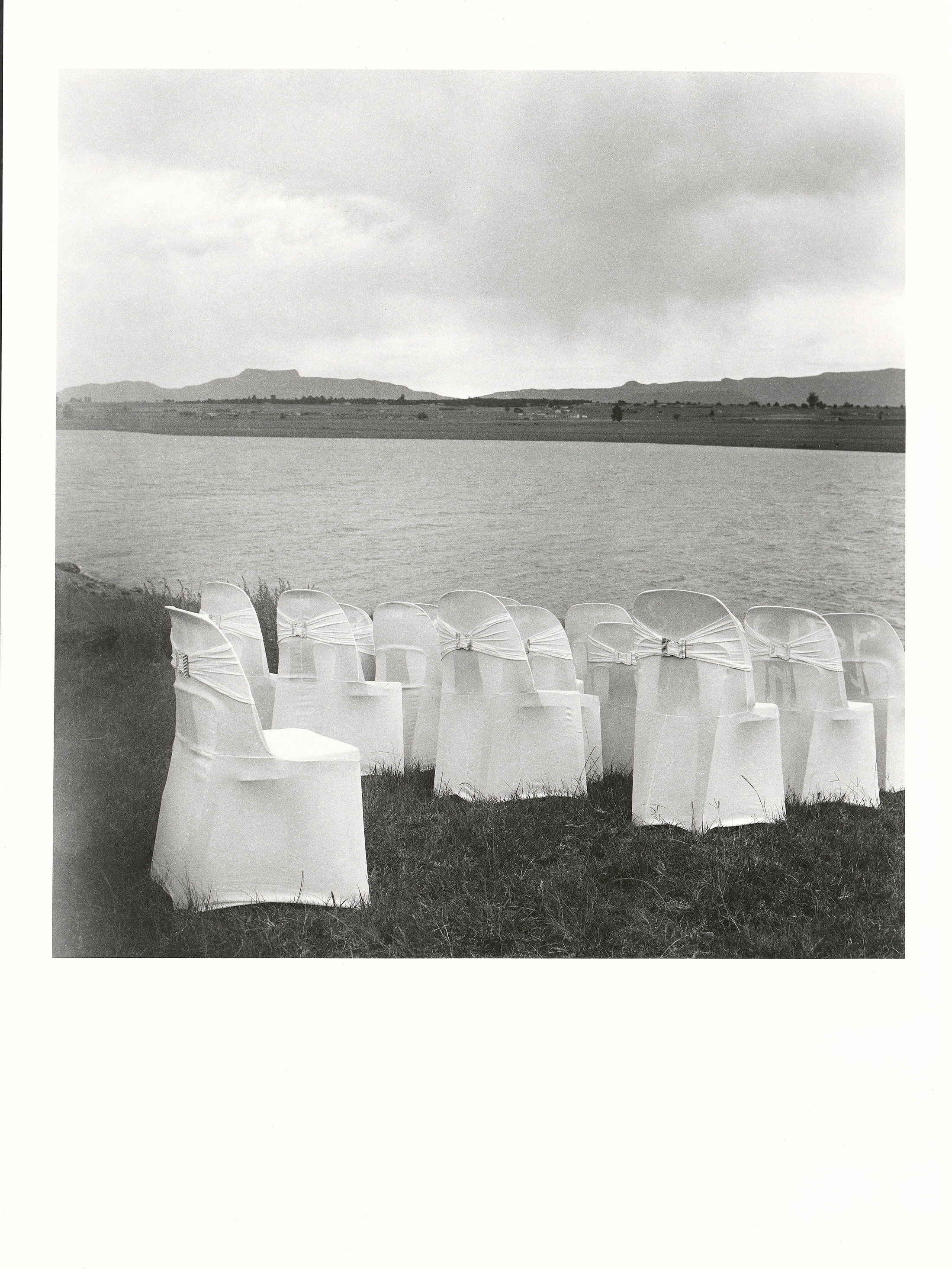 Sabelo Mlangeni’s ‘Ukufika kwengilosi. The coming of an angel’, a black and white photograph depicting chairs covered in cloth overlooking a river.
