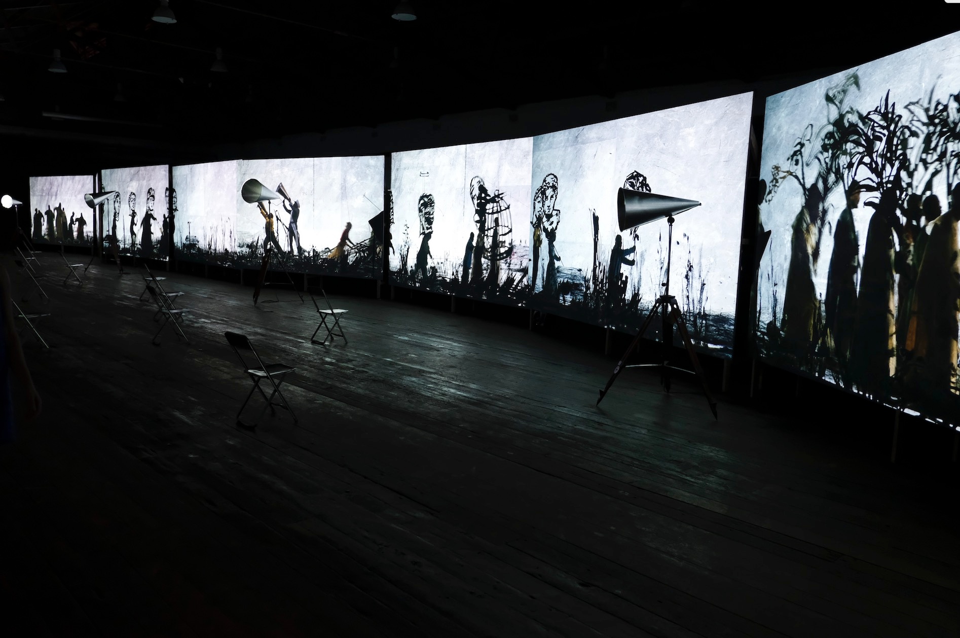 Installation photograph of William Kentridge’s video installation ‘More Sweetly play the Dance’ from the series of offsite exhibitions ‘Crossing Night / Hacer Noche’ in Oaxaca, Mexico. At the back, multiple videos are projected onto a gallery wall in a darkened room, with 4 metal megaphones on stands arranged at intervals in front of it. At the front, collapsable metal chairs are arranged in rows with lots of intervening space.
