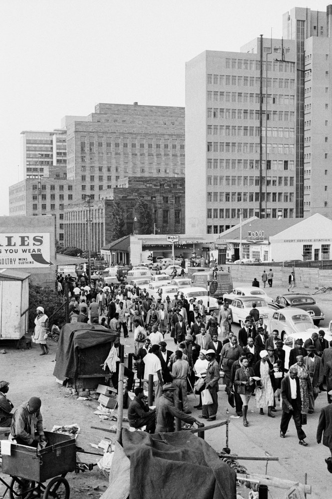 David Goldblatt's monochrome photograph 'Evening exodus on West Street: Blacks going for trains in Soweto, Whites in their cars to the suburbs, Johannesburg' shows two lines of individuals in a cityscape moving in opposite directions. On the left, pedestrians walking towards the photographer. On the right, a line of cars moving away from the photographer.
