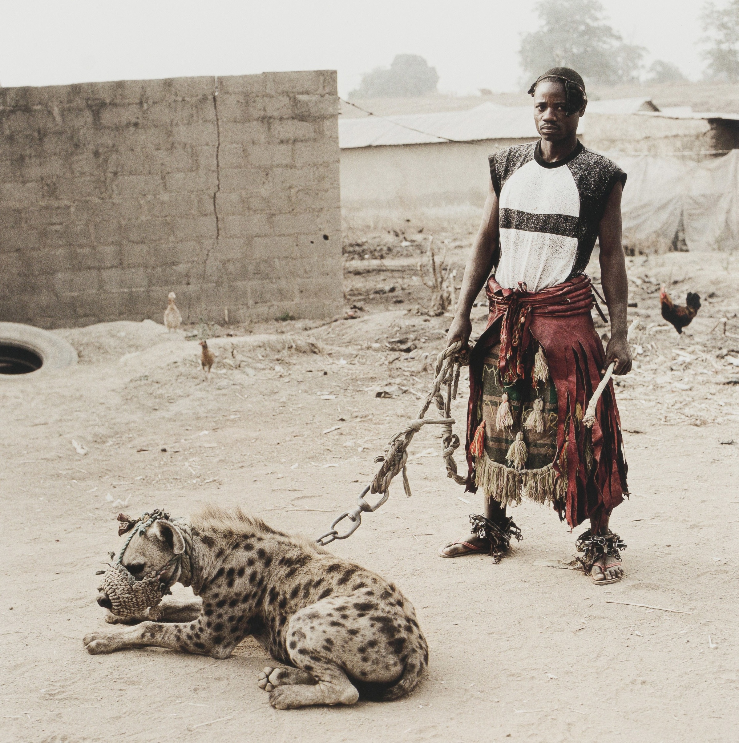 Pieter Hugo’s photograph ‘Mallam Mantari Lamal with Mainasara, Abuja, Nigeria, 2005’ depicts an individual holding a hyena on a leash.

