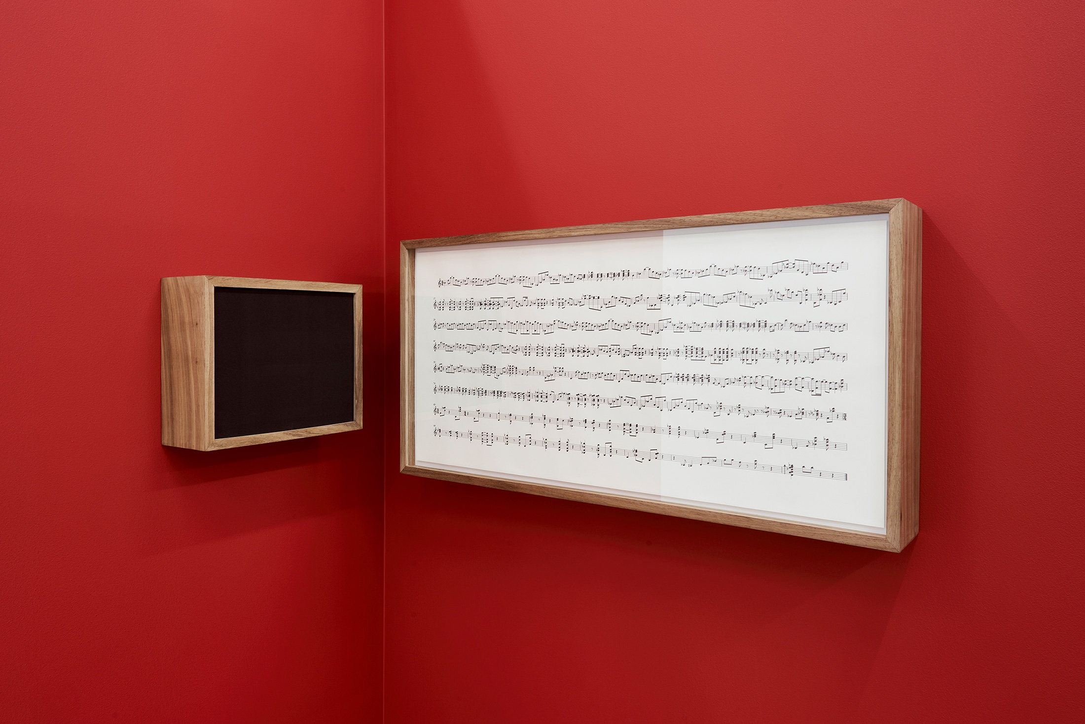 Installation photograph that shows Iñaki Bonillas’ wall-mounted installation ‘The Return to the Origin 6’ in the corner of two red movable gallery walls. On the left is a wood-framed speaker box and on the right, a wood-framed musical score.
