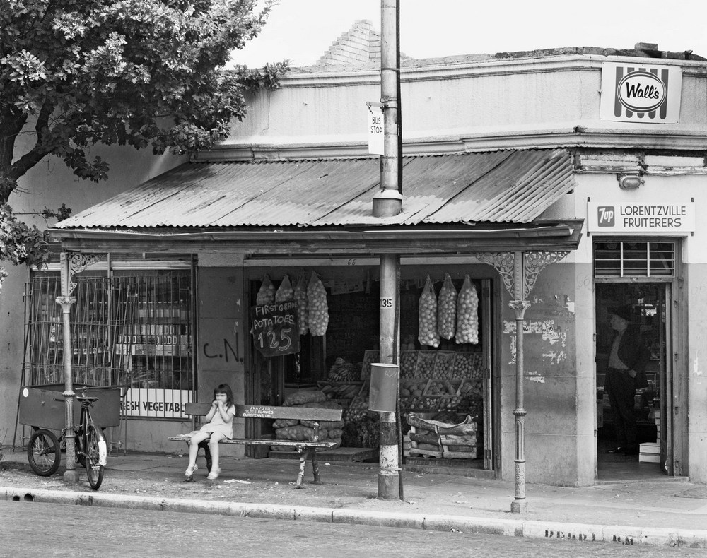 David Goldblatt's black-and-white photograph 'Bus stop, Derby Road, Lorentzville, Johannesburg. December' shows a young girl sitting on a bench. Behind her is a storefront selling fruits and vegetables.
