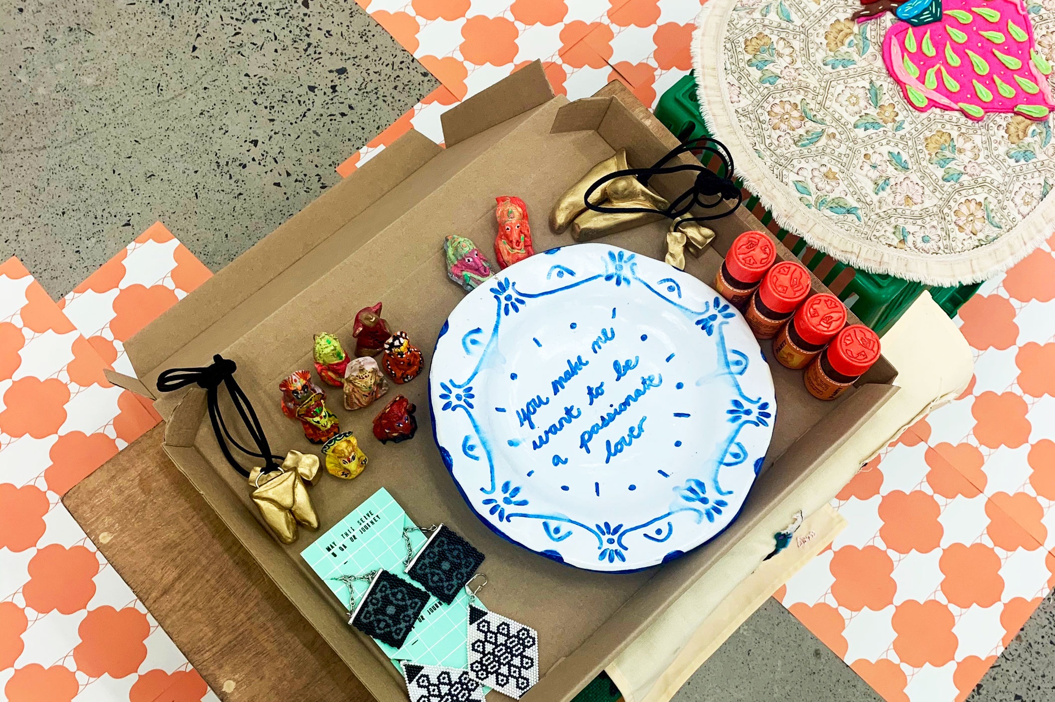 Photograph of ephemera from ‘The Kutti Bazaar,’ the Kutti Collective’s online pop-up in collaboration A4’s Proto~ museum shop. In the centre, a small table supports a flat cardboard box holding various small art objects, with sheets of patterned paper lining the concrete floor below it. In the middle, Githan Coopoo’s ceramic plate reads “you make me want to be a a passionate lover.” On the right, Tazme Pillay’s aromatherapy bottles ‘PARTY’. On the left, Youlendree Appasamy’s ‘Goth Kolam earrings’ and ‘Minimal Kolam earrings’. 
