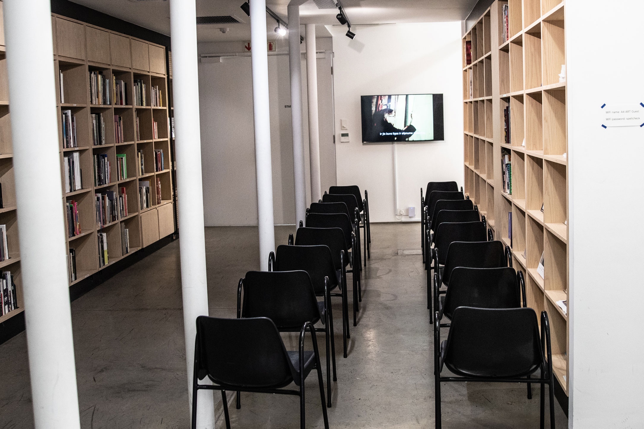 Event photograph from the screening of Jonas Mekas' film 'Zefiro Torna' in A4 Arts Foundation Library. At the back, a wall-mounted screen plays the film. At the front, two rows of plastic chairs. At the left and right, the library bookshelves.
