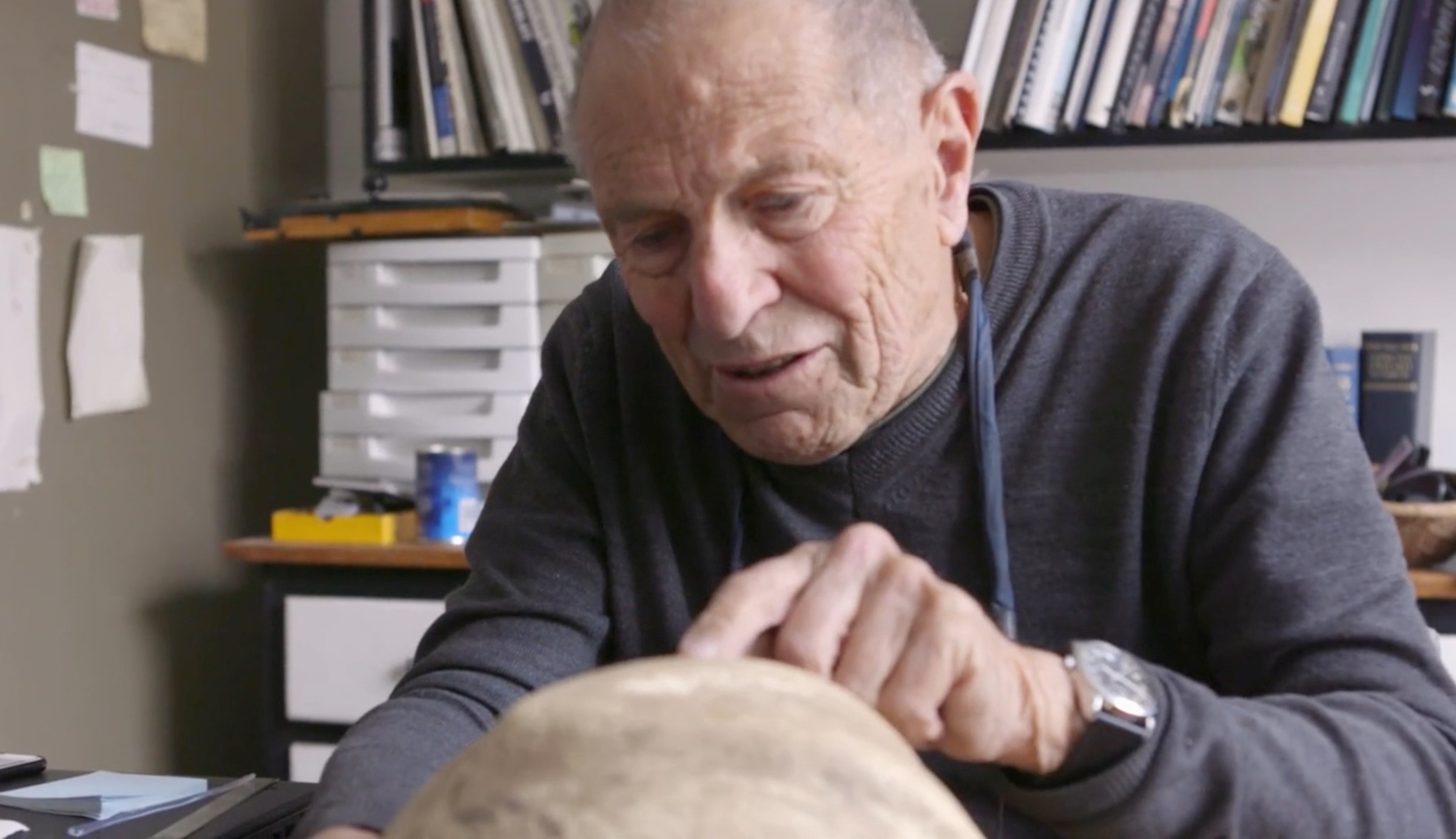 Process photograph from the making of Daniel Zimbler's documentary on David Goldblatt that shows Goldblatt pointing at an object, with a desk and shelf at the back.
