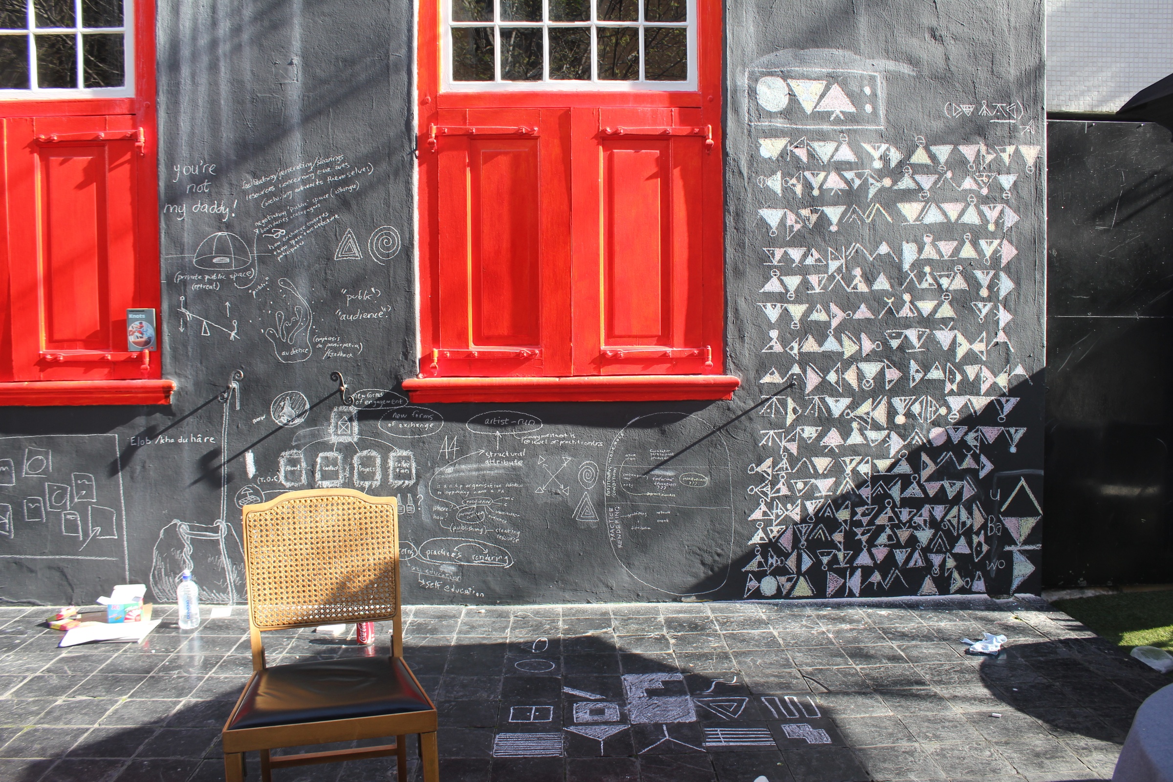 Process photograph from A4’s ‘Stoep Residency’ at the Association of Visual Arts (AVA) in Cape Town. At the back, the AVA’s wall is covered in chalk drawings. At the front, a wooden chair sits next to chalk drawings on the AVA’s veranda.
