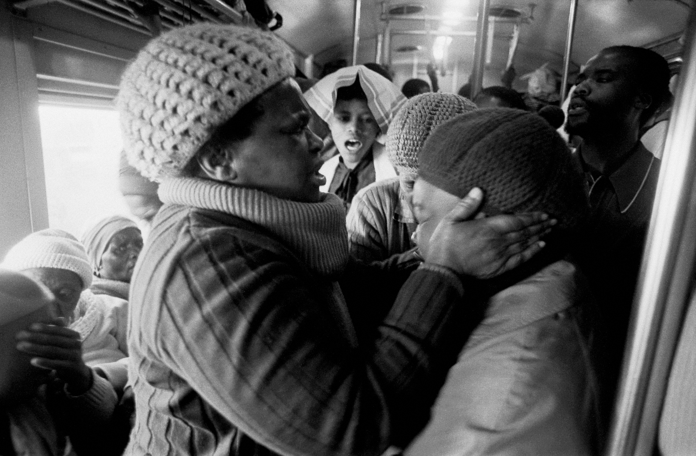 Santu Mofokeng's monochrome photographic print 'Supplication, Johannesburg–Soweto Line' shows two individuals standing inside a railway vehicle, one holding the head of the other with both hands.
