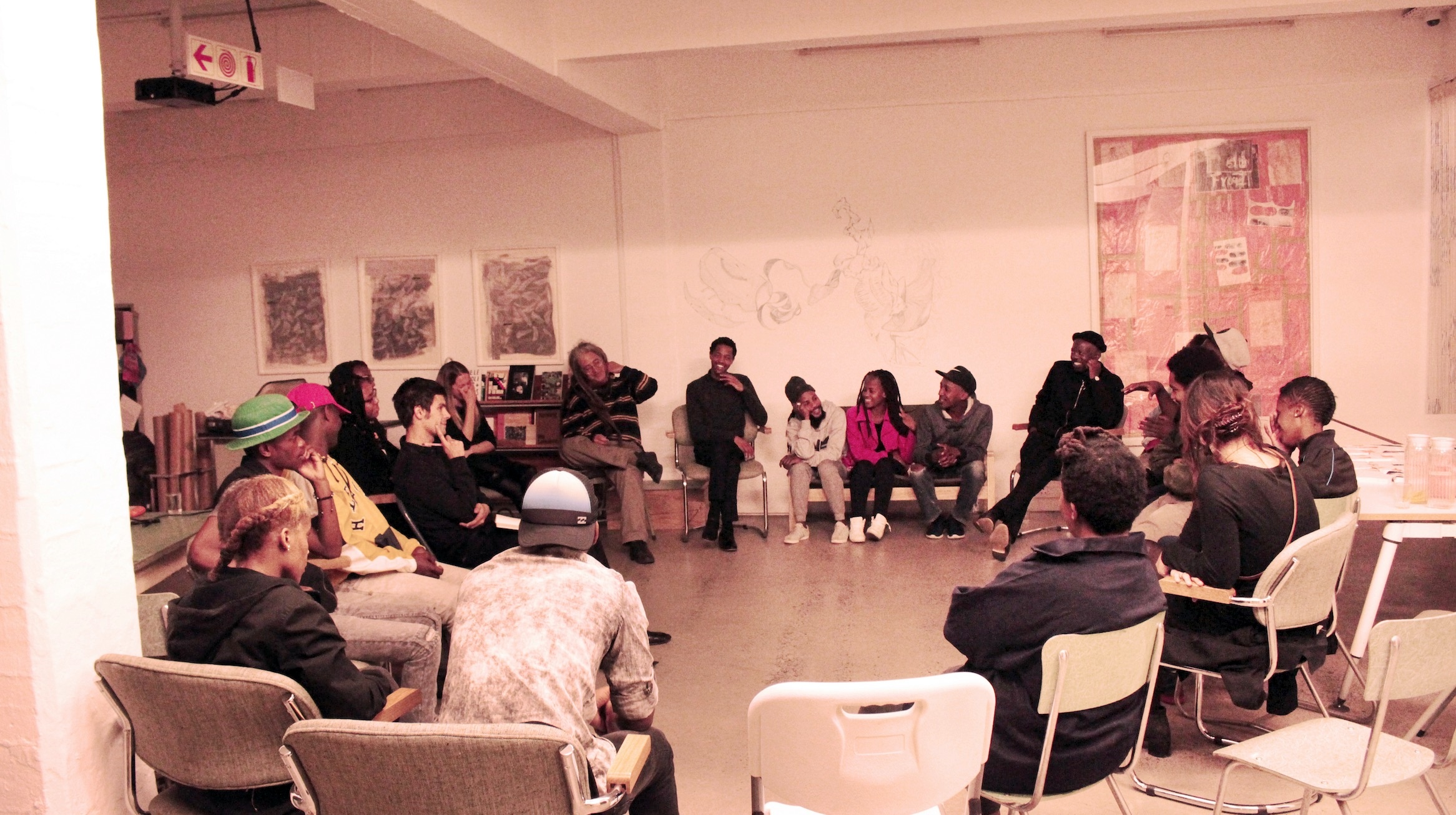 Monochrome event photograph from the ‘Creating in Circles’ exchange on A4’s ground floor. At the front, the facilitator Eugene Paramoer seated along with participants in a circular formation. At the back, Nolan Oswald Dennis’ mixed media drawings ‘abangenamalihlaba IV-VI’ and Moshekwa Langa’s mixed media work ‘Ramothibedi’ are mounted on the wall, with a drawing on the wall between them.
