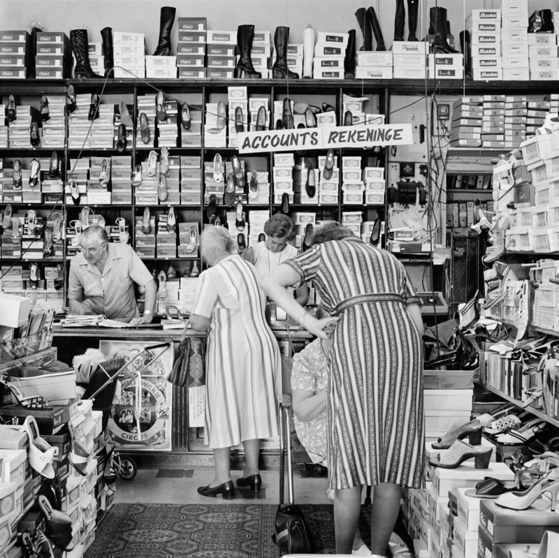 David Goldblatt's black-and-white photograph 'In a family outfitting store, Boksburg' shows women gathered in a store, surrounded by shoes.
