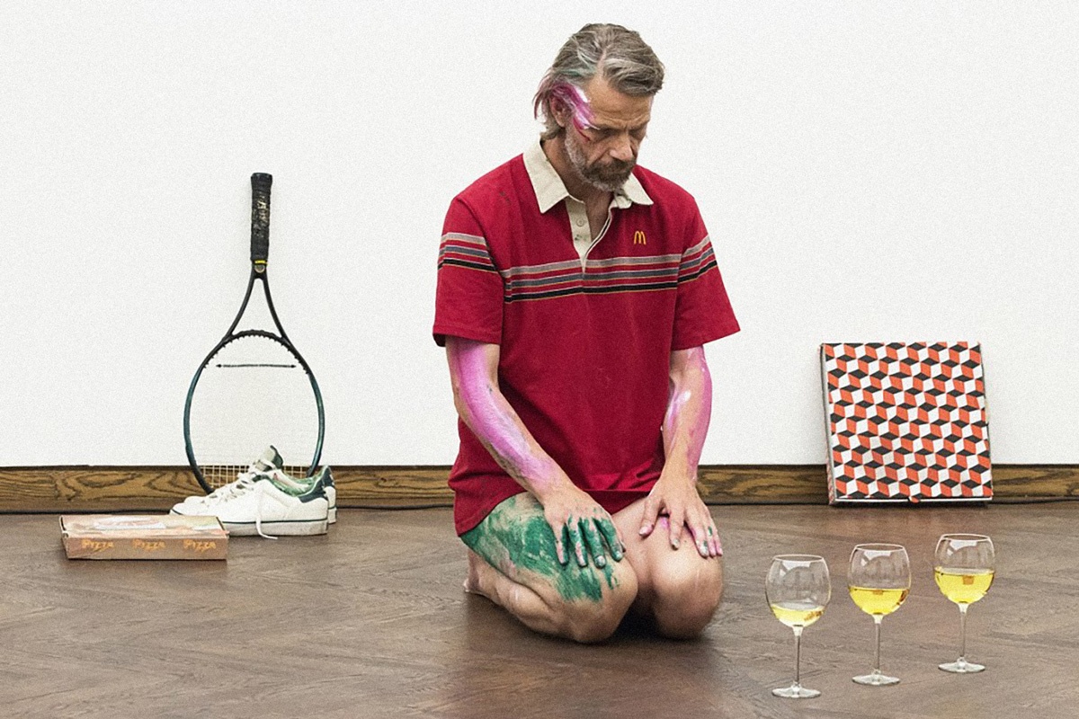 Event photograph from Mårten Spångberg’s solo dance ‘Digital Technology’ on A4’s top floor. In the middle, the artist is seated on the floor wearing a branded shirt, with paint marks on his legs, arms and face. On the left, a tennis racket and shoes lean against the wall behind him, along with a pizza box. On the right, three wine glasses filled with yellow liquid sit on the floor, and panel with a three dimensional cubic pattern rests against the wall behind them.
