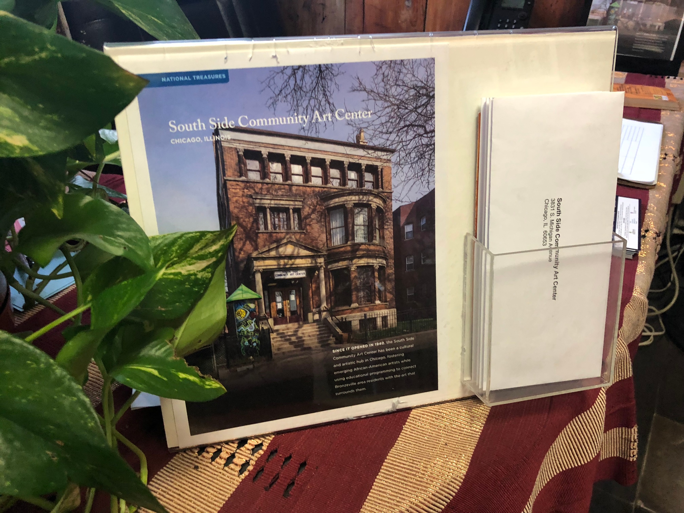Process photograph from the 2018 offsite rendition of Curatorial Exchange with Art Institute Chicago. An acrylic document stand holds a photograph about the South Side Community Art Center in Chicago, Illinois, along with pre-addressed envelopes to the Center.
