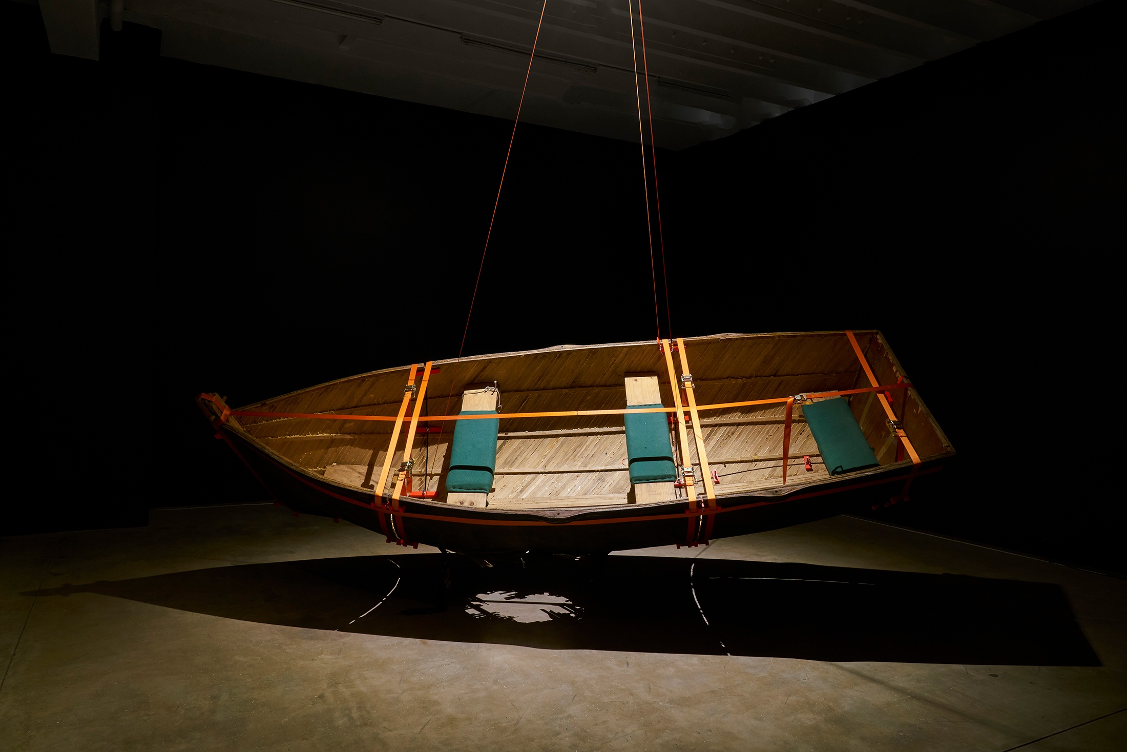 Installation photograph from the ‘Risk’ exhibition in A4’s Gallery. In the middle Gimberg Nerf’s mixed media installation ‘Escape to Robben Island (Angasi Nkosi Angasi Nkosi)’ consists of a wooden rowing boat suspended from the ceiling, tilted towards the viewer.
