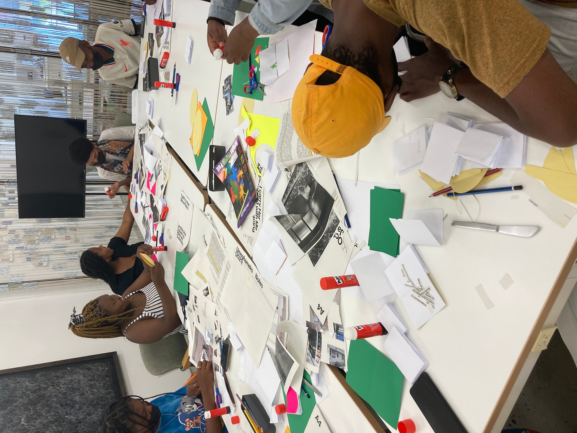 Event photograph from the ‘Lalela x A4' exchange during the ‘Tell It to the Mountains’ exhibition in A4’s Gallery. Lalela students are seated around white laminate tables with cutting and pasting tools in A4’s Reading Room.
