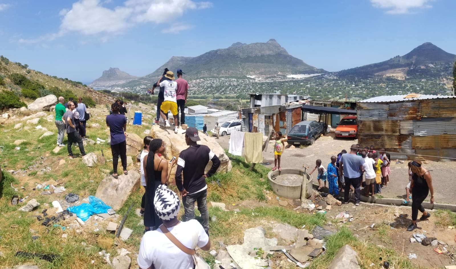 Process photograph from the offsite ‘Ghetto Relay - Lalela experiment’ exchange. At the front, various participants stand on the edge of the ‘Imizamo Yethu’ township in Hout Bay, with houses made of corrugated iron plating visible on the right. At the back, Table Mountain is visible in the distance.
