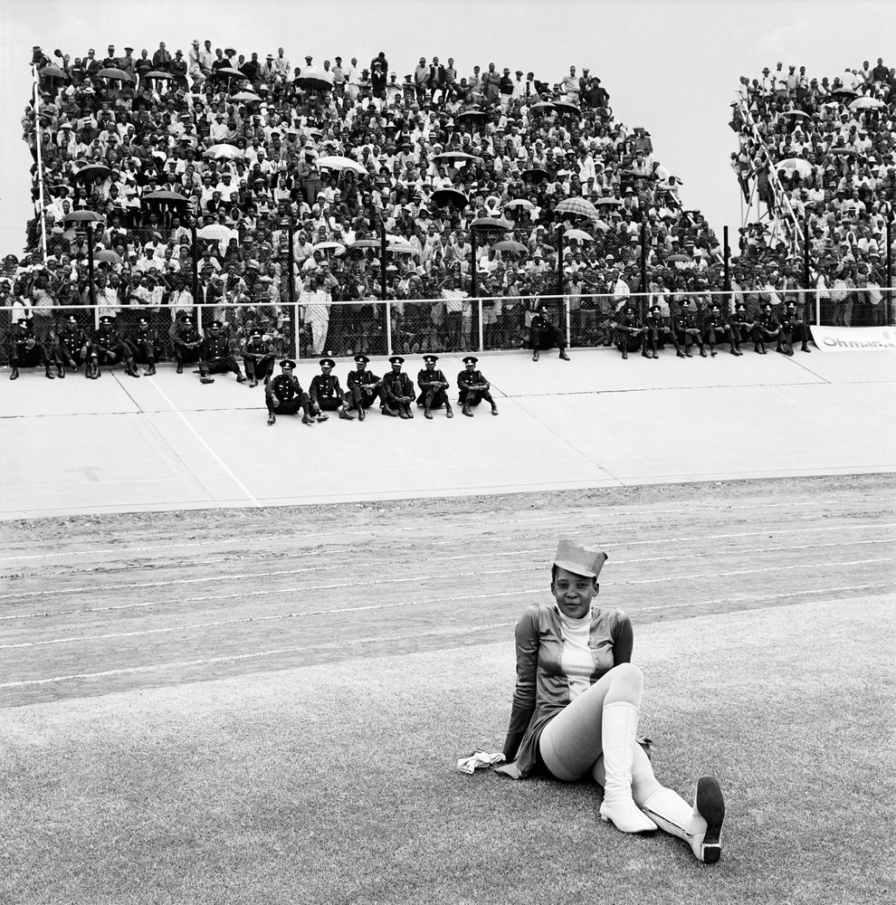David Goldblatt's black-and-white photograph 'Drum majorette, Cup final, Orlando Stadium, Soweto. 1972' shows a drum majorette sitting on a field with a crowd in the background.
