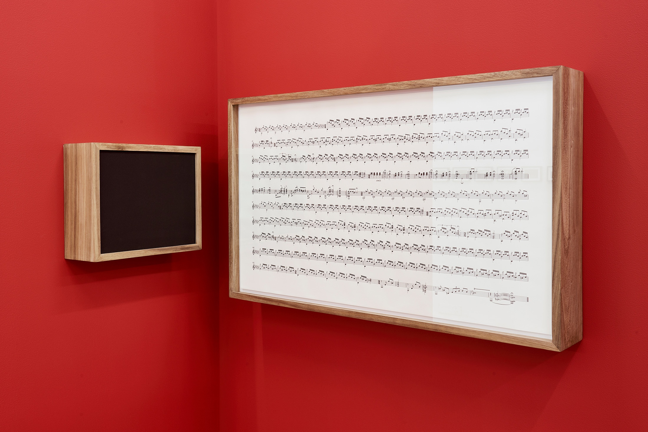 Installation photograph that shows Iñaki Bonillas’ wall-mounted installation ‘The Return to the Origin 8’ in the corner of two red movable gallery walls. On the left is a wood-framed speaker box and on the right, a wood-framed musical score.
