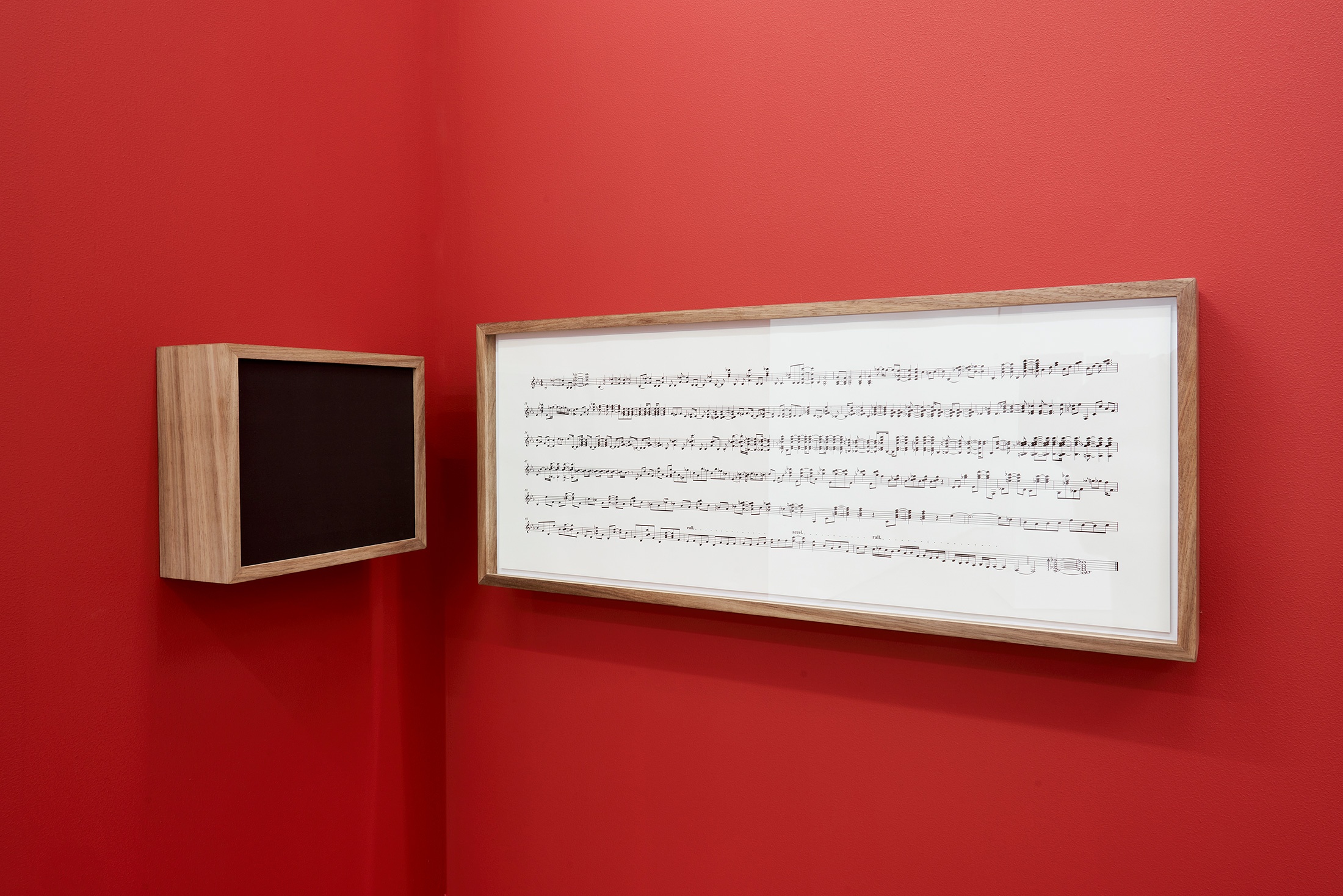 Installation photograph that shows Iñaki Bonillas’ wall-mounted installation ‘The Return to the Origin 5’ in the corner of two red movable gallery walls. On the left is a wood-framed speaker box and on the right, a wood-framed musical score.
